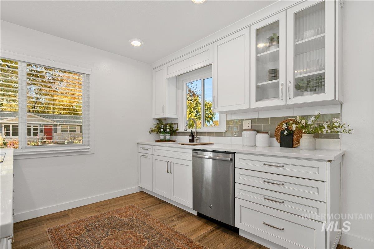 Kitchen featuring white cabinets, glass insert cabinets, stainless steel dishwasher, backsplash, and dark wood-style floors