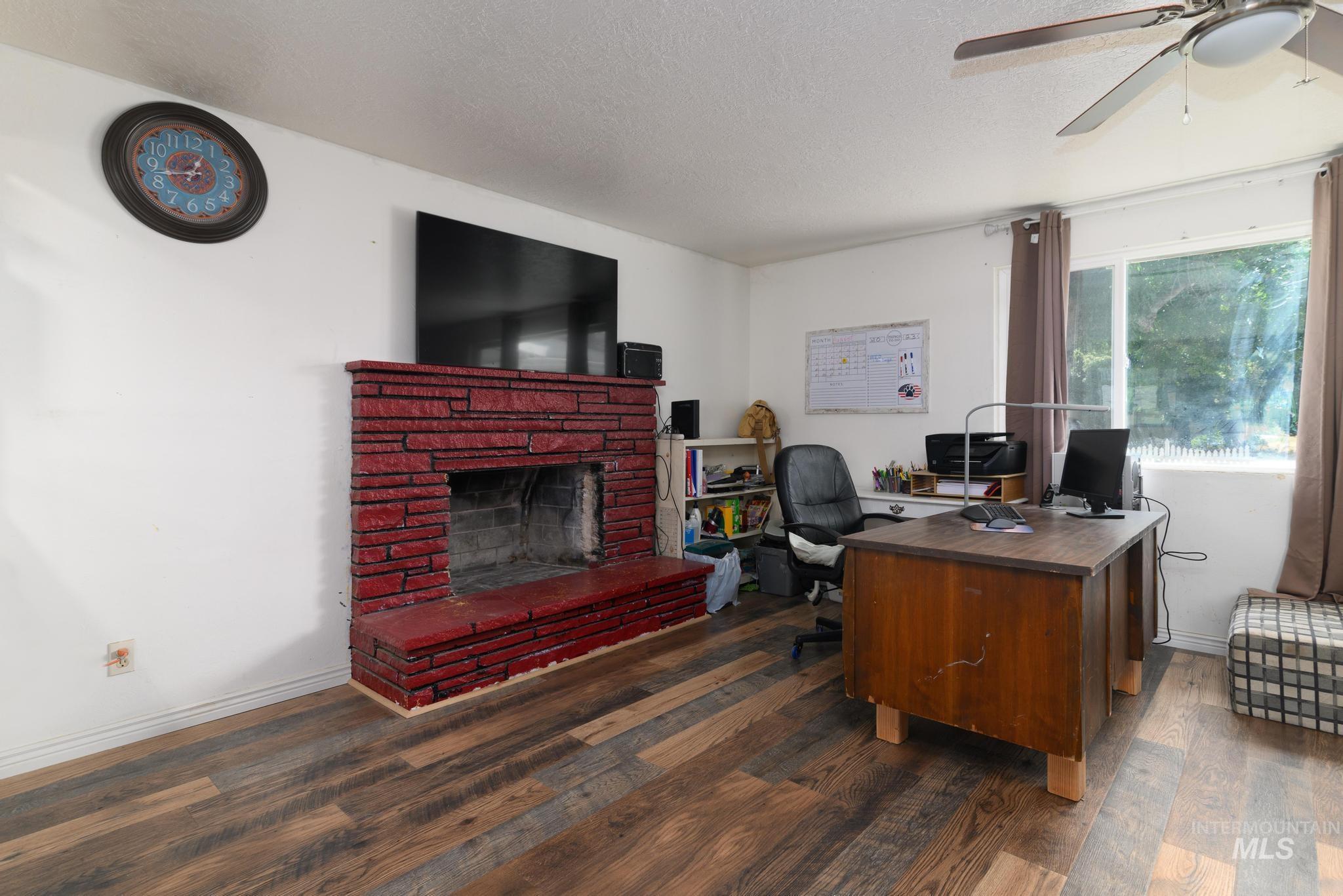 Office area with a brick fireplace, a textured ceiling, dark wood-type flooring, and a ceiling fan