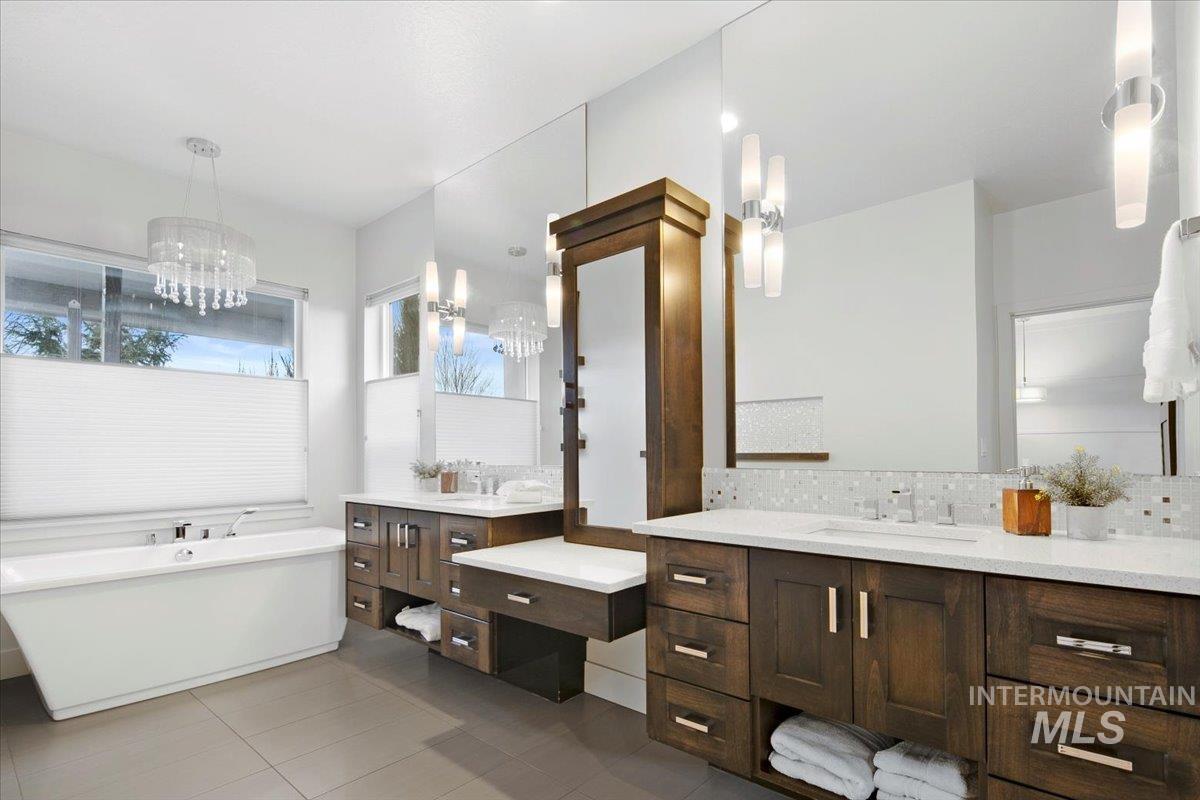Full bath featuring light tile patterned floors, backsplash, two vanities, a freestanding tub, and a chandelier