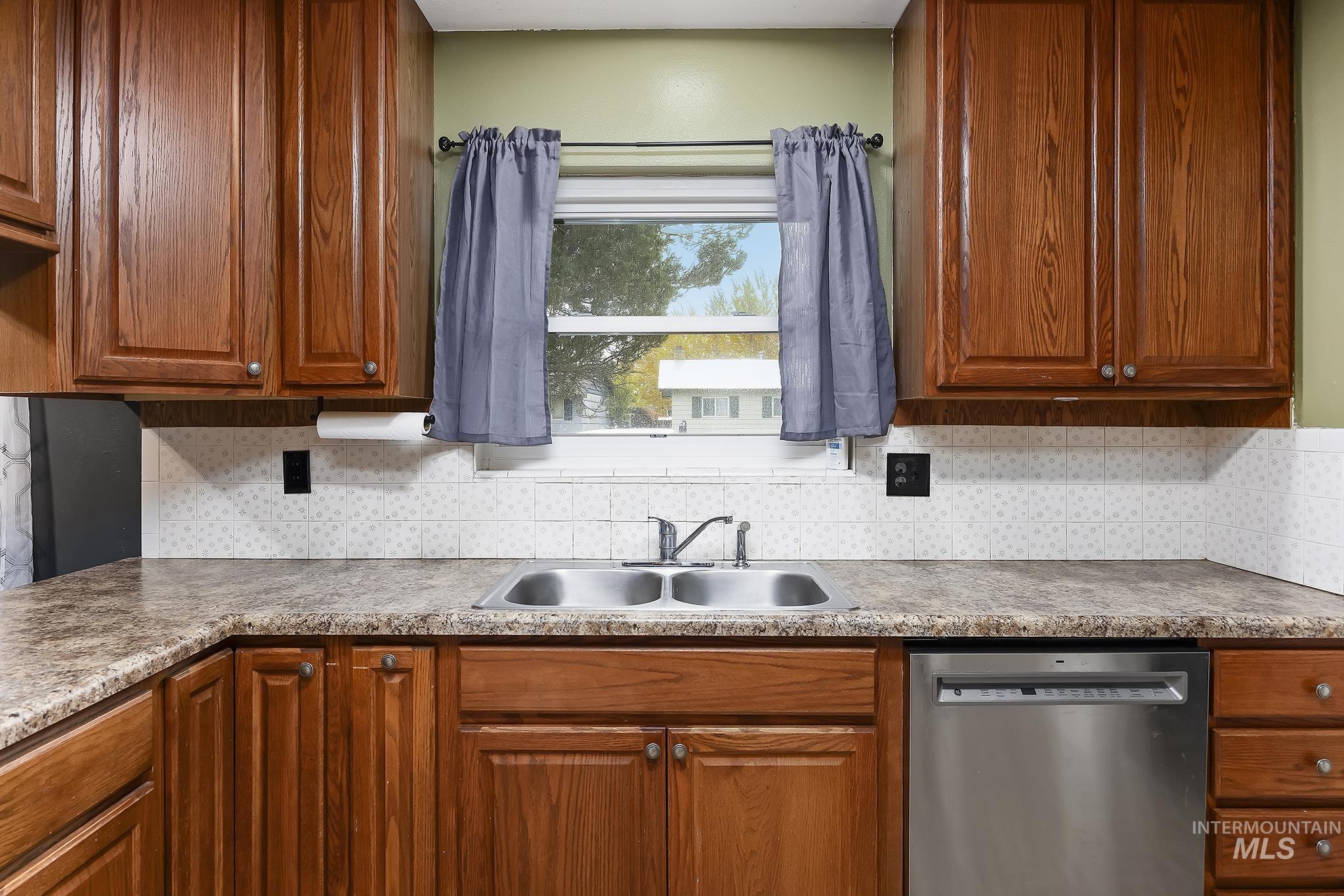 Kitchen featuring stainless steel dishwasher, brown cabinetry, decorative backsplash, and light countertops