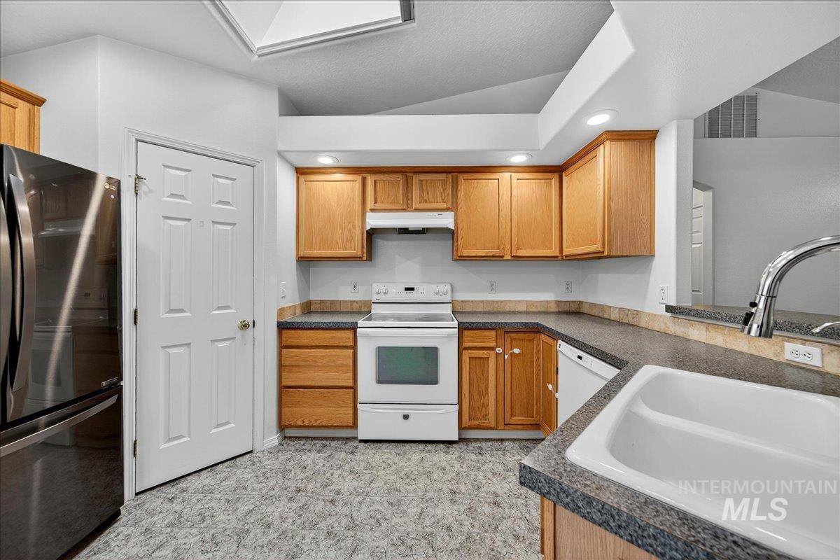 Kitchen featuring white appliances, dark countertops, recessed lighting, brown cabinetry, and under cabinet range hood