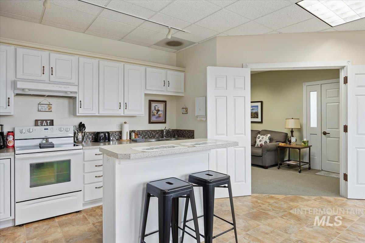Kitchen featuring white electric range, a kitchen breakfast bar, light countertops, white cabinetry, and a drop ceiling