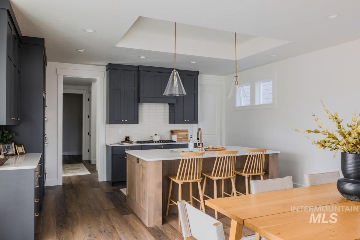 Kitchen with a raised ceiling, a center island with sink, decorative backsplash, dark wood-style flooring, and hanging light fixtures