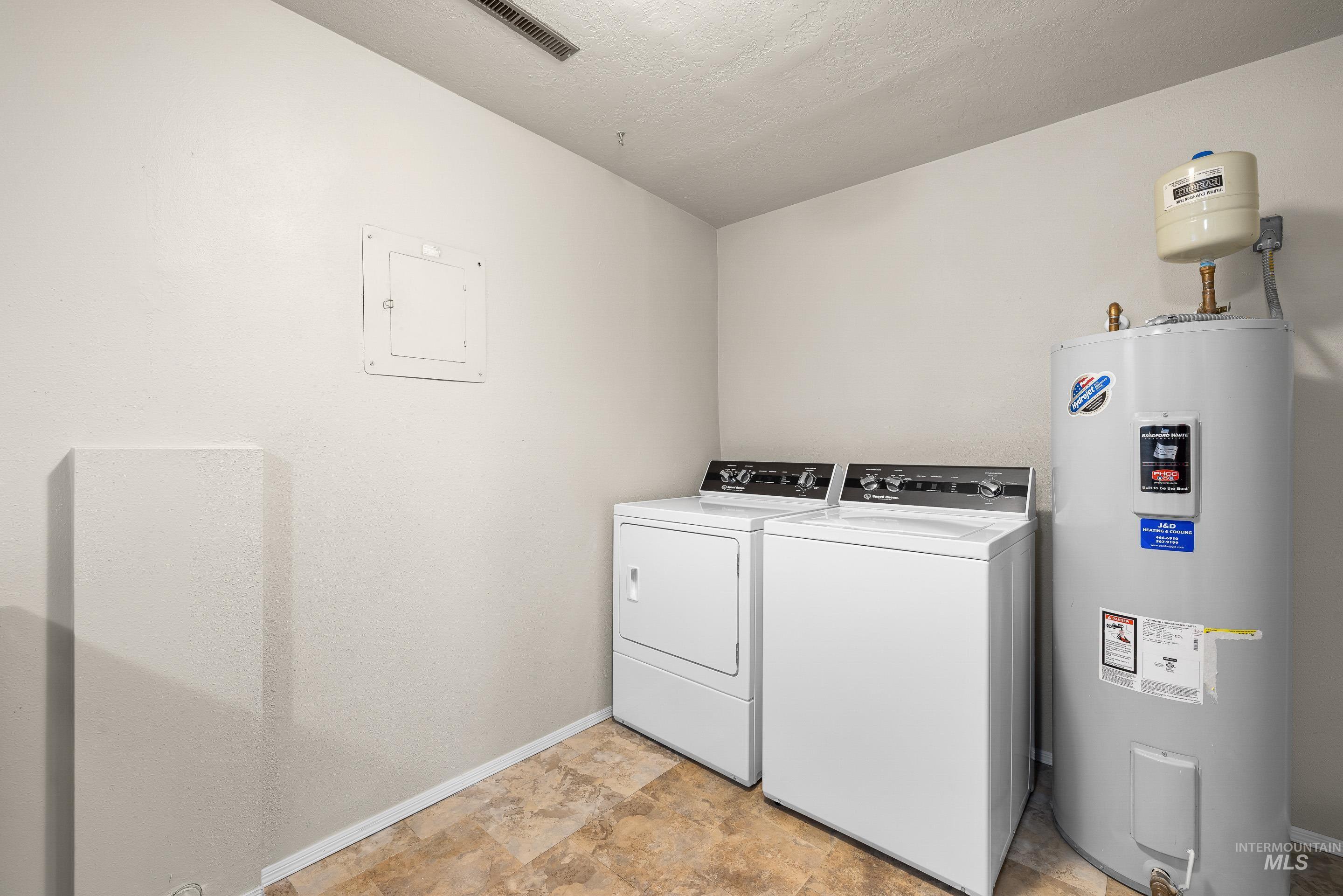 Washroom with water heater, electric panel, washer and dryer, and stone finish flooring