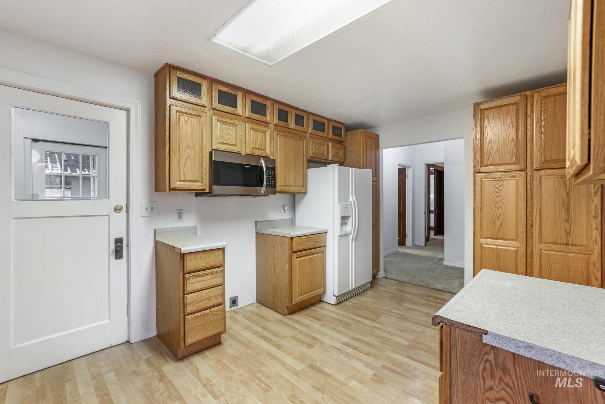 Kitchen featuring white refrigerator with ice dispenser, light wood finished floors, light countertops, glass insert cabinets, and stainless steel microwave