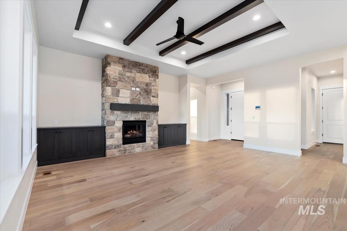 Unfurnished living room featuring ceiling fan, beamed ceiling, light wood-style floors, a raised ceiling, and a stone fireplace