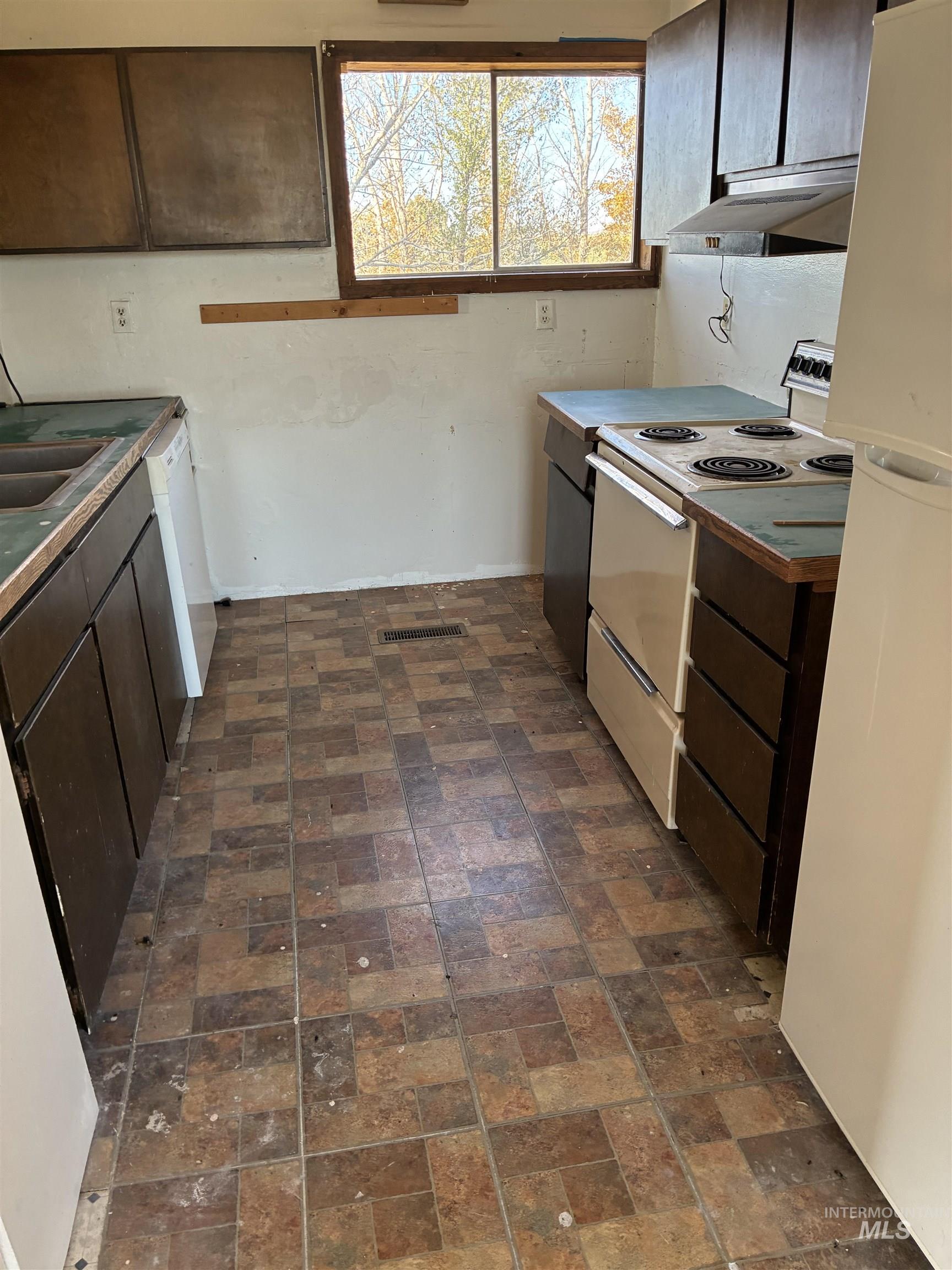 Kitchen featuring dark brown cabinetry, white appliances, under cabinet range hood, and brick patterned floors