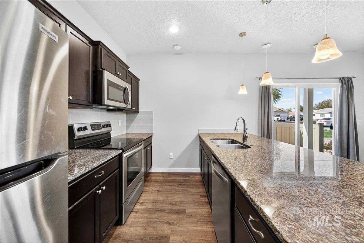 Kitchen featuring appliances with stainless steel finishes, dark wood-type flooring, light stone counters, hanging light fixtures, and a textured ceiling