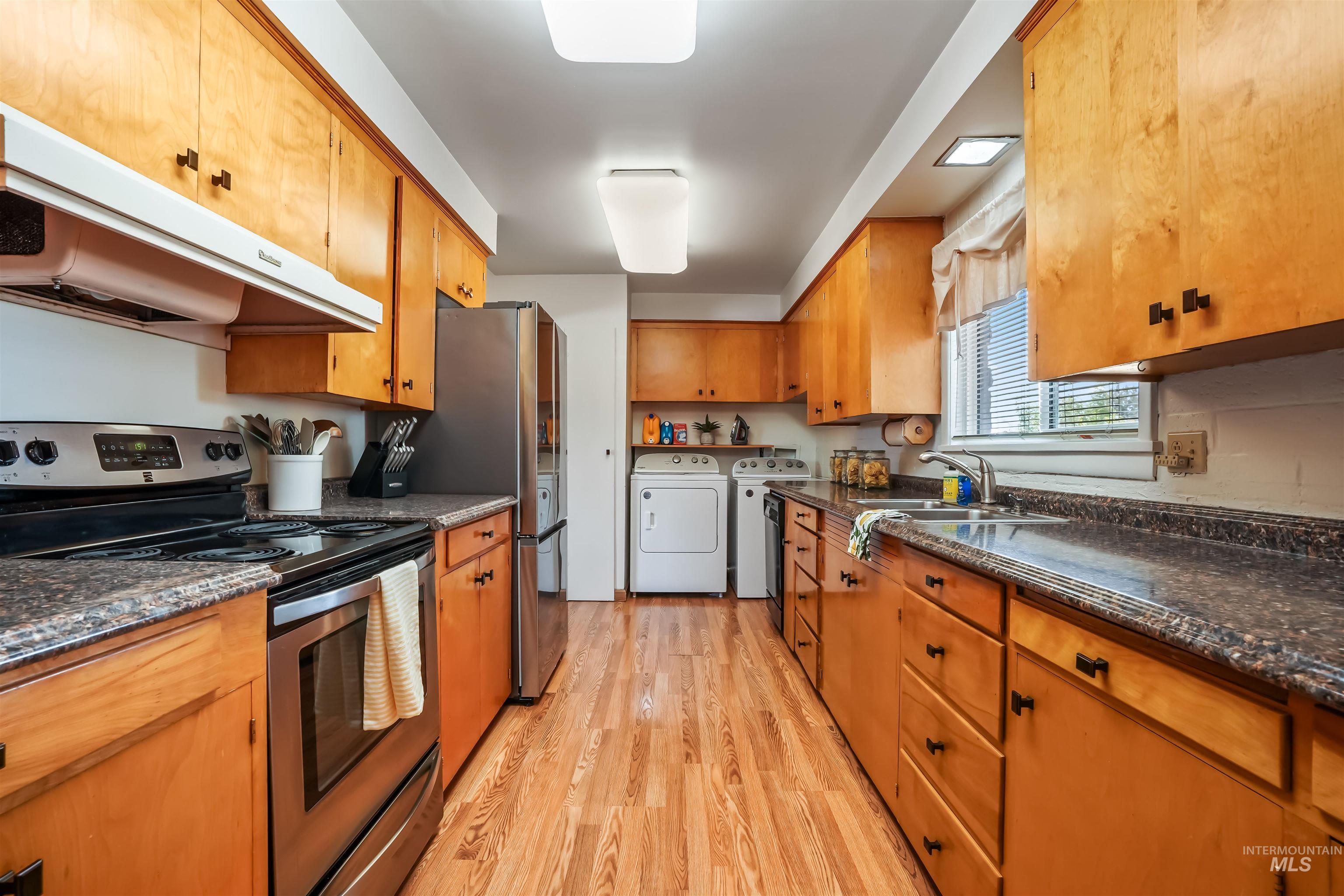 Kitchen with stainless steel electric stove, light wood-style floors, ventilation hood, brown cabinetry, and washer and dryer