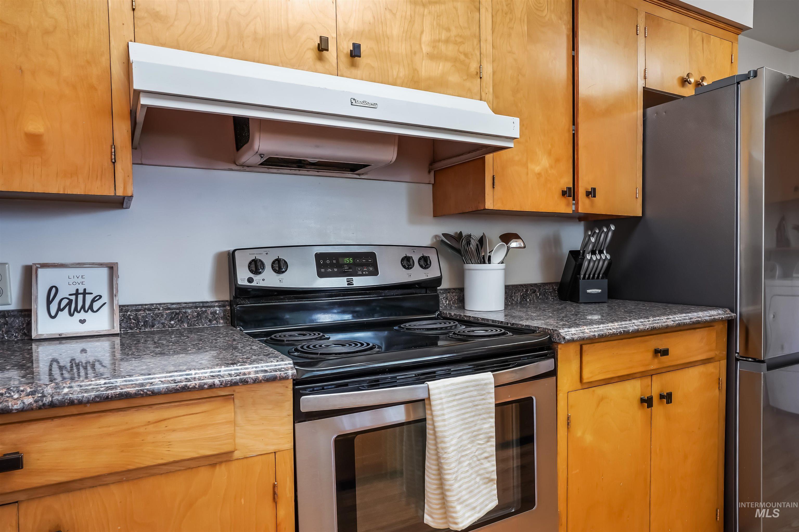 Kitchen with exhaust hood, stainless steel electric stove, brown cabinetry, and dark stone countertops