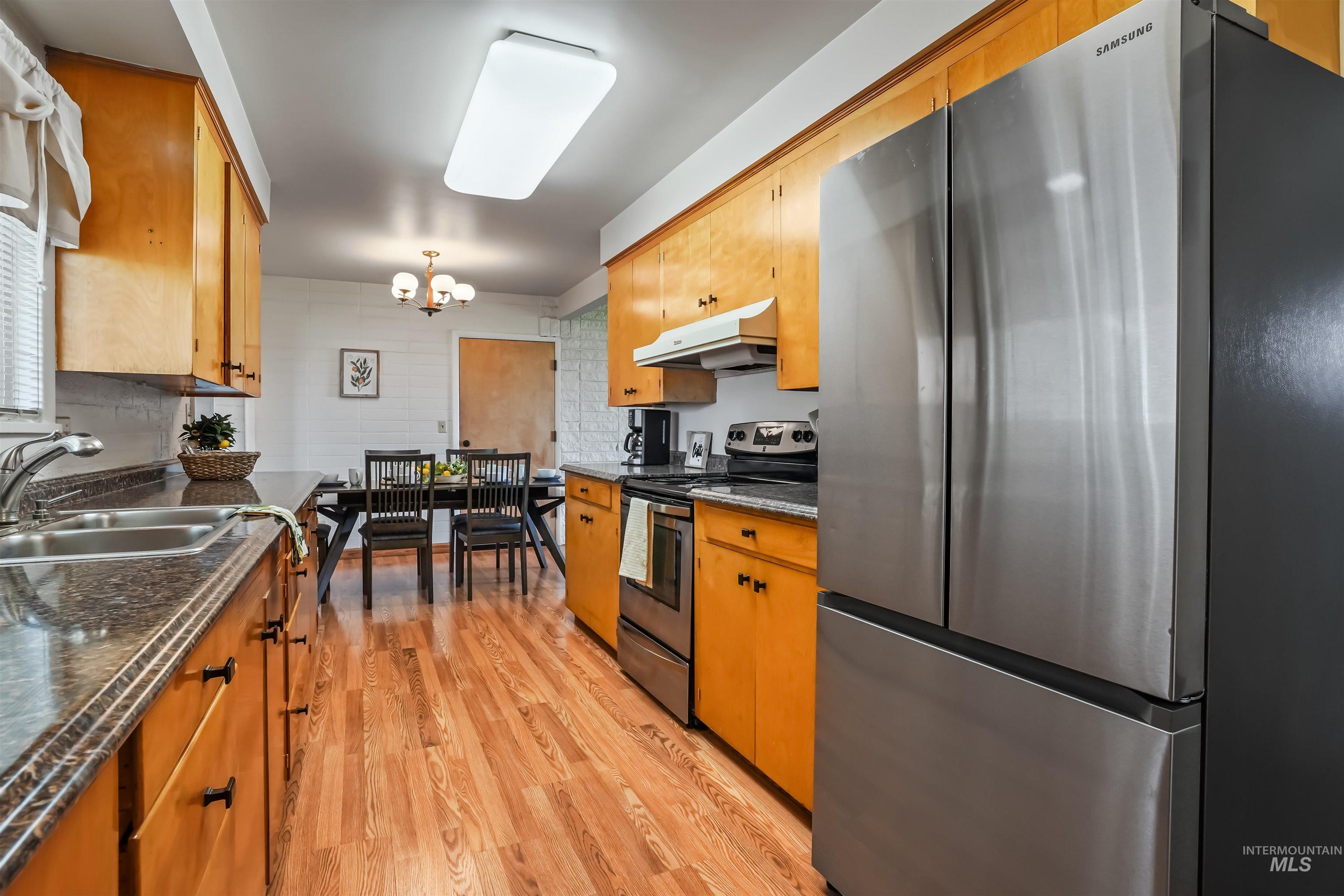 Kitchen featuring stainless steel appliances, under cabinet range hood, light wood finished floors, a chandelier, and dark stone counters