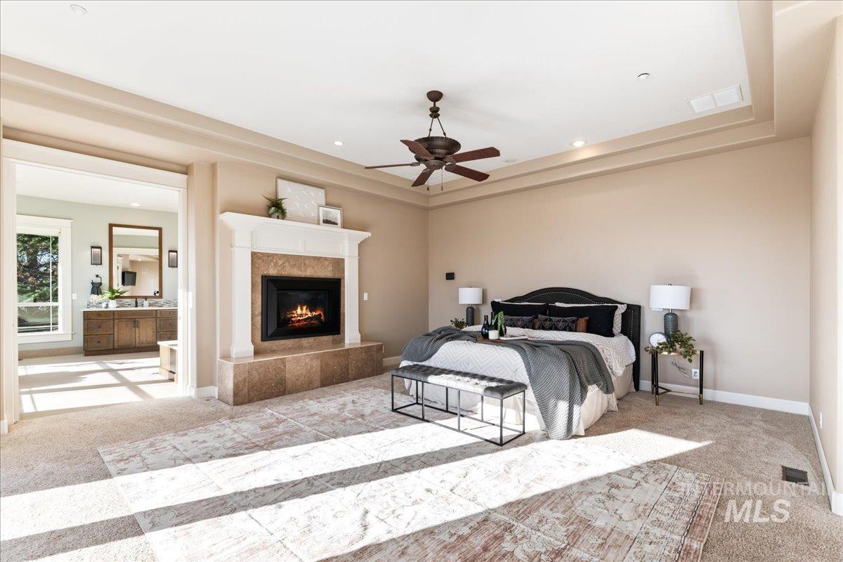 Carpeted bedroom with a tray ceiling, a ceiling fan, a lit fireplace, ensuite bath, and recessed lighting