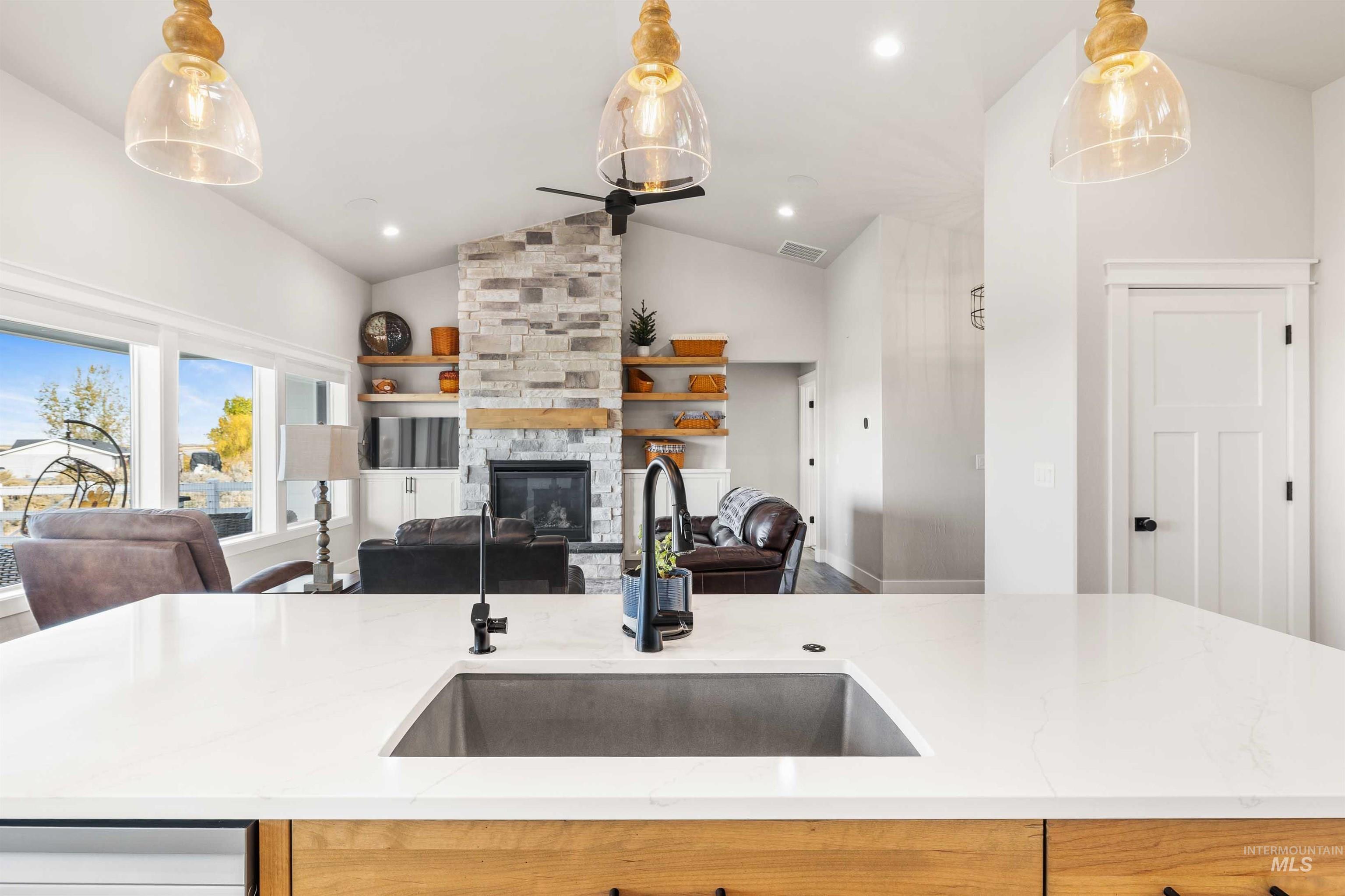 Kitchen featuring lofted ceiling, hanging light fixtures, light stone counters, an island with sink, and a stone fireplace