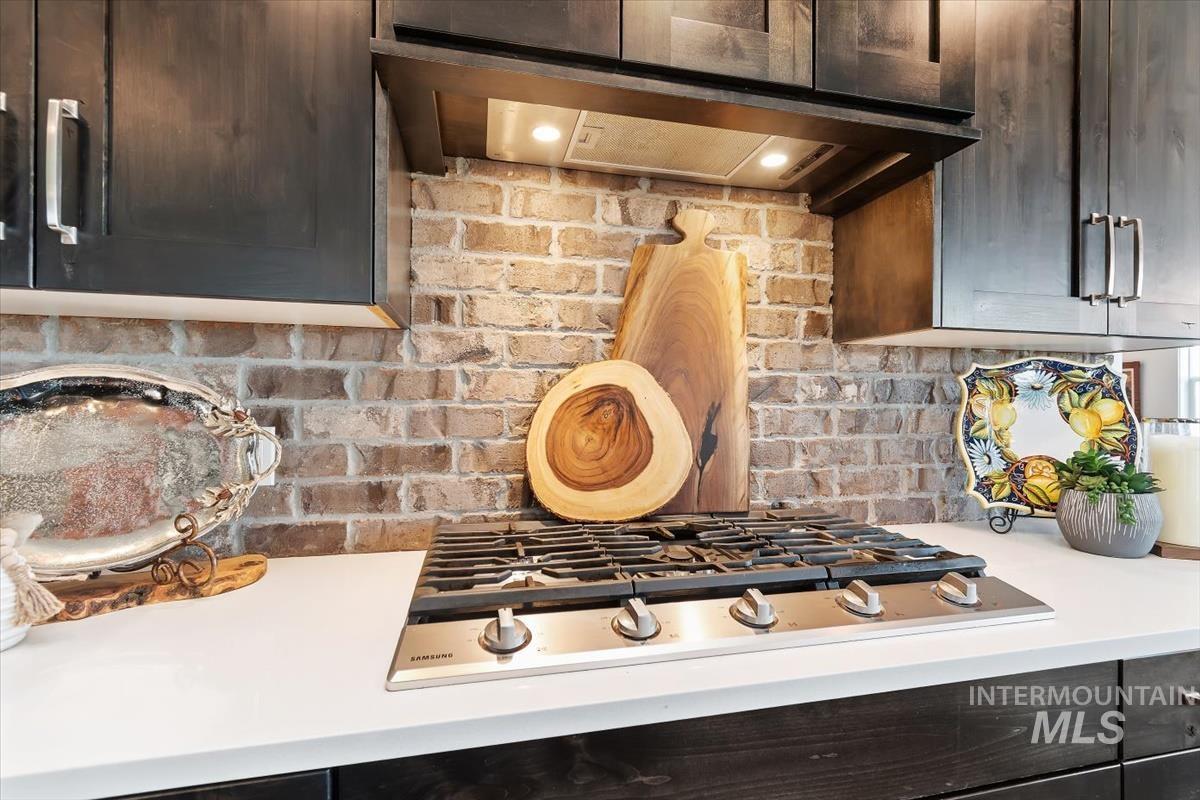 Kitchen view of extractor fan, stainless steel gas cooktop, dark brown cabinets, backsplash, and light stone countertops