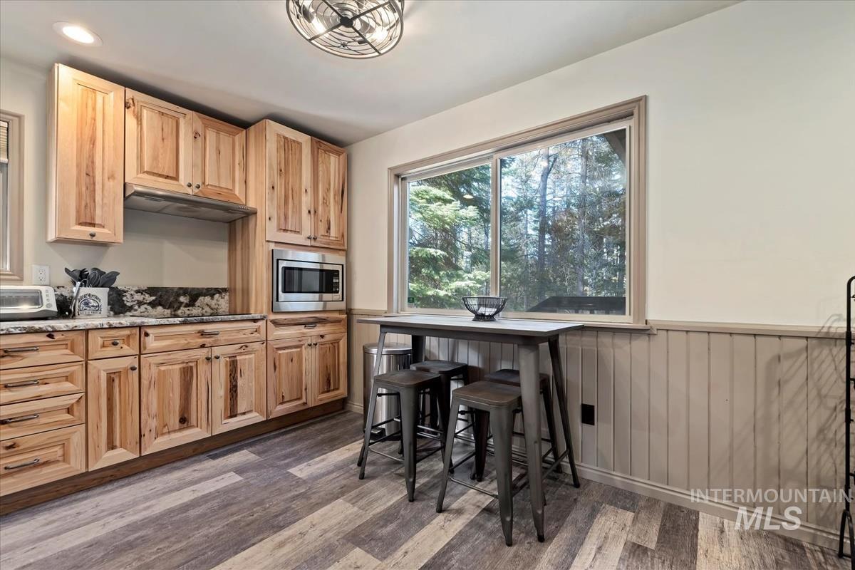 Kitchen with a wainscoted wall, dark wood-type flooring, stainless steel microwave, dark stone countertops, and under cabinet range hood