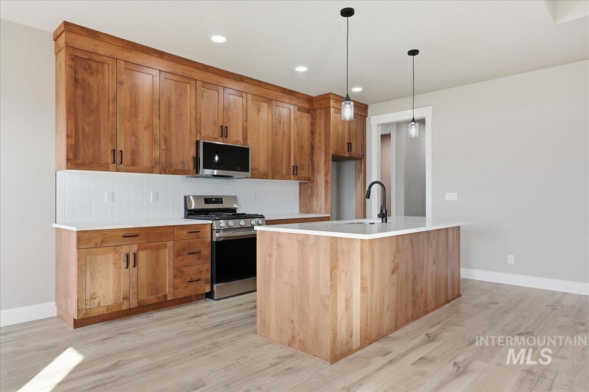 Kitchen with stainless steel appliances, brown cabinetry, backsplash, a kitchen island with sink, and decorative light fixtures