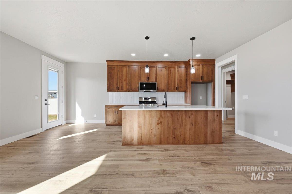 Kitchen with brown cabinetry, a kitchen island with sink, pendant lighting, appliances with stainless steel finishes, and light wood-type flooring