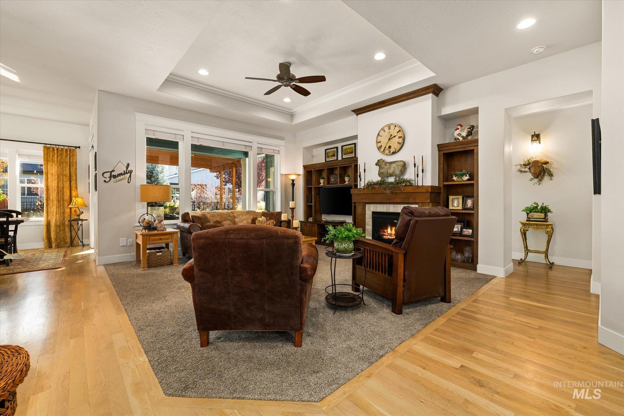 Living room featuring light wood-type flooring, ceiling fan, a tiled fireplace, recessed lighting, and a tray ceiling