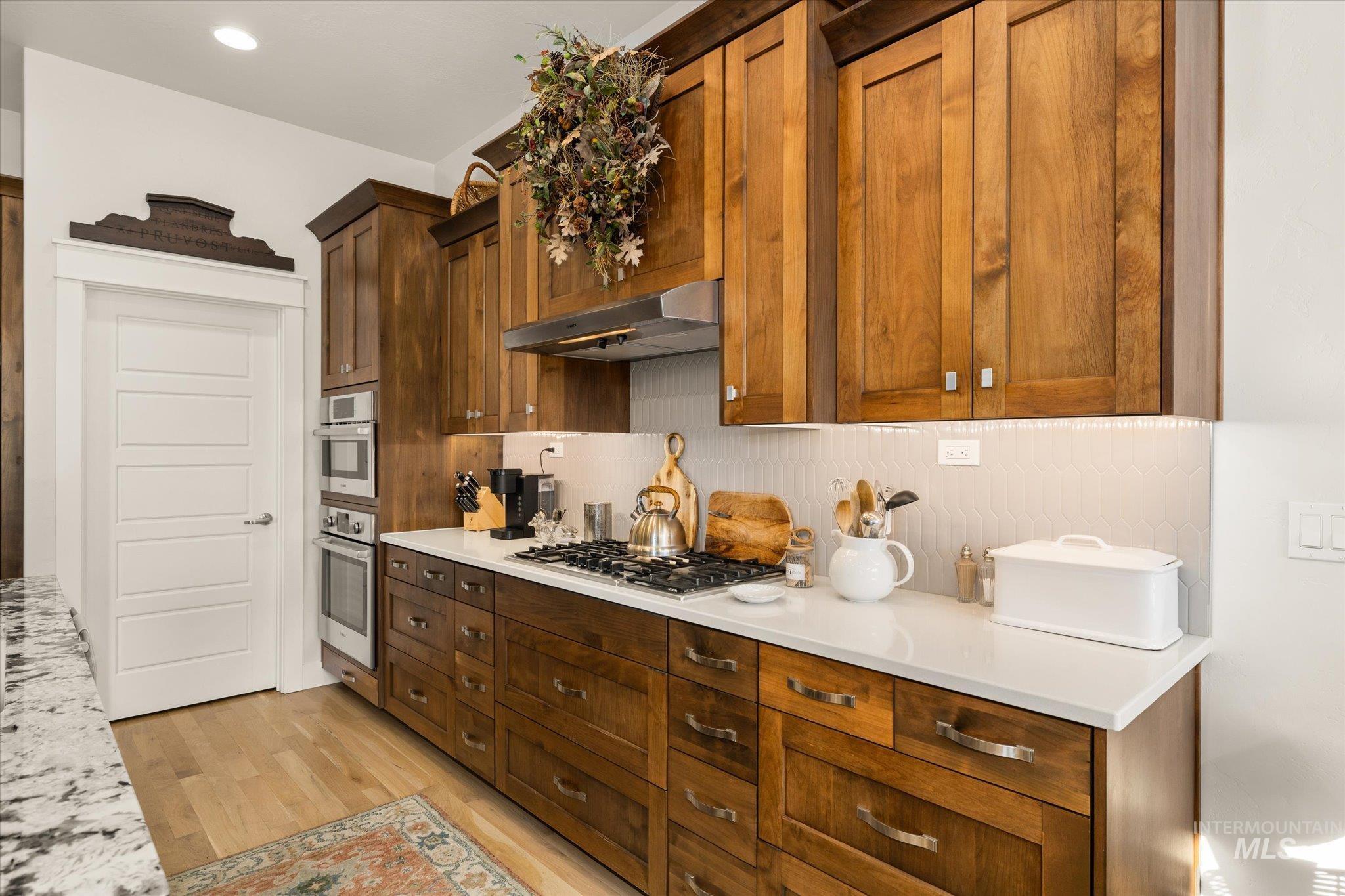 Kitchen with under cabinet range hood, light wood-style floors, light stone countertops, appliances with stainless steel finishes, and tasteful backsplash