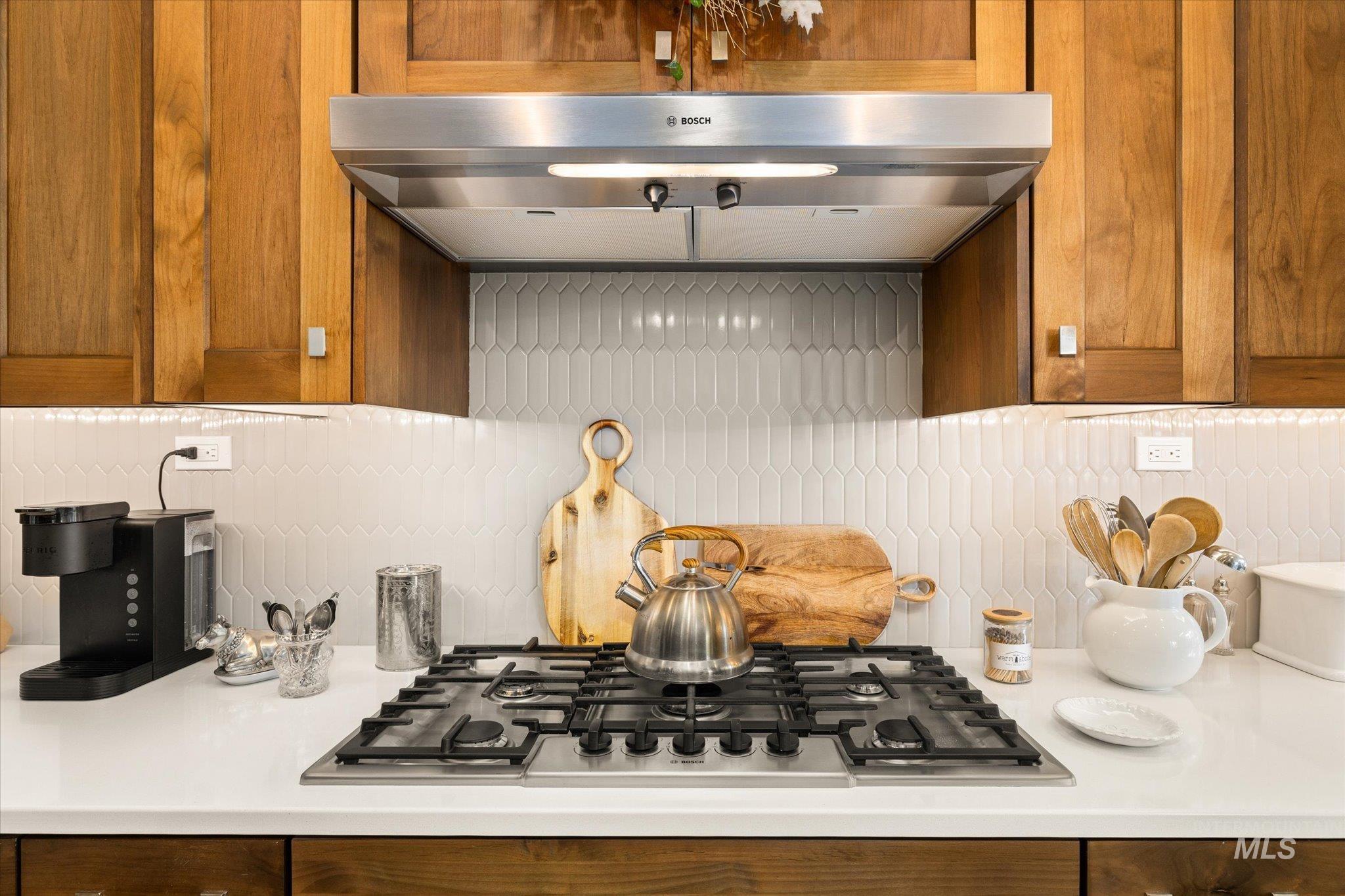 Kitchen featuring under cabinet range hood, decorative backsplash, stainless steel gas stovetop, and brown cabinetry