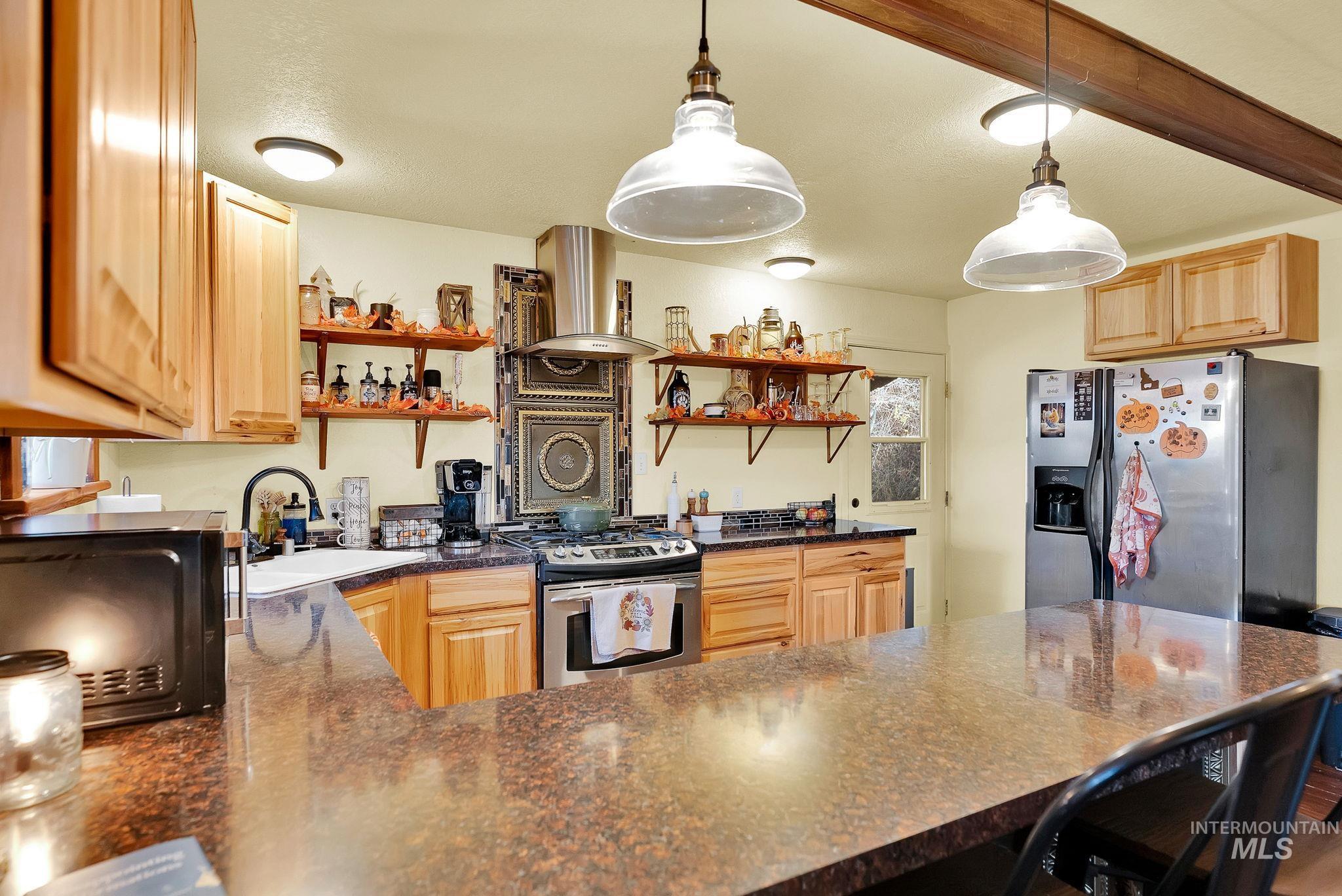 Kitchen with dark stone counters, stainless steel appliances, hanging light fixtures, open shelves, and ventilation hood