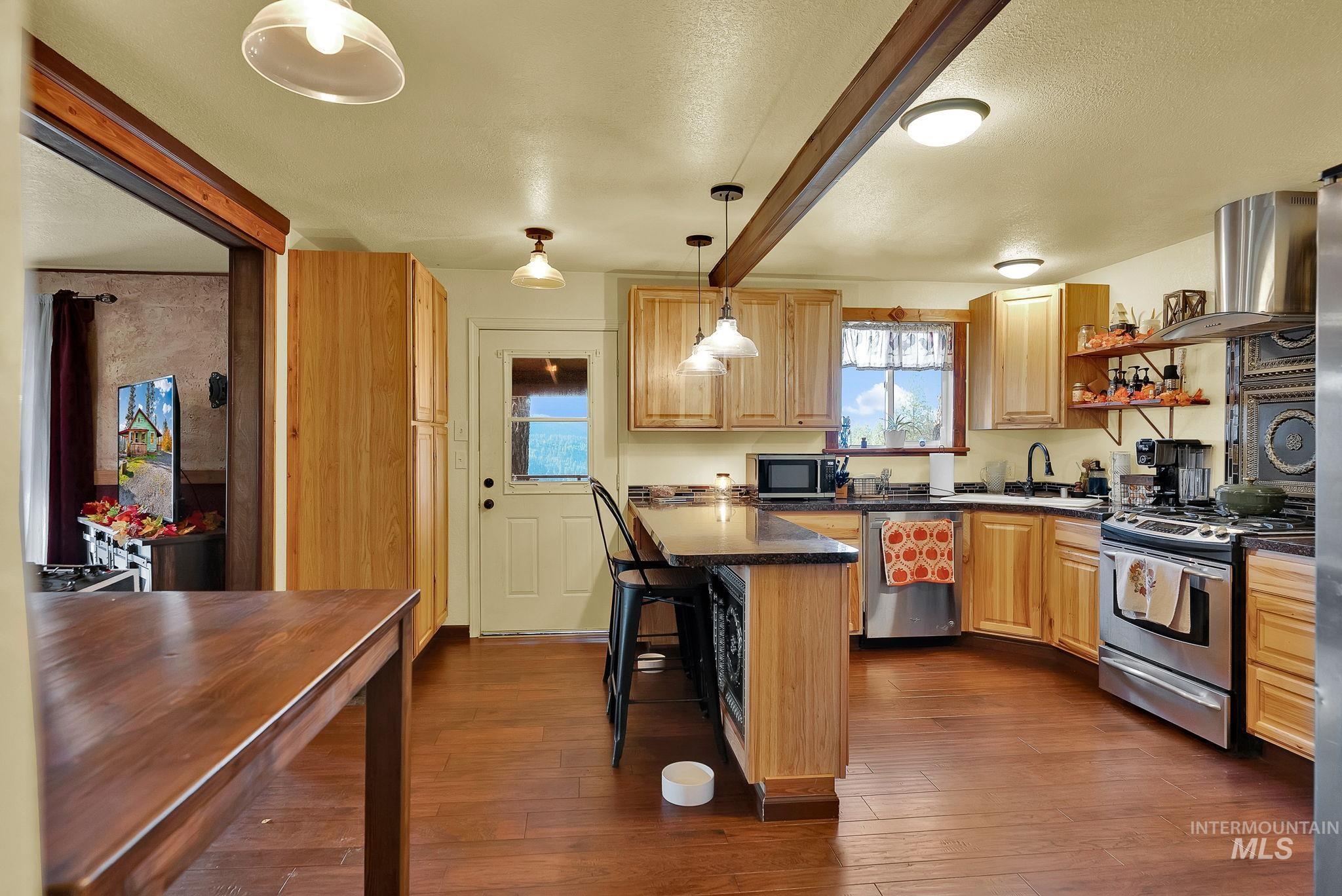 Kitchen featuring beam ceiling, stainless steel appliances, a kitchen bar, decorative light fixtures, and dark wood-type flooring
