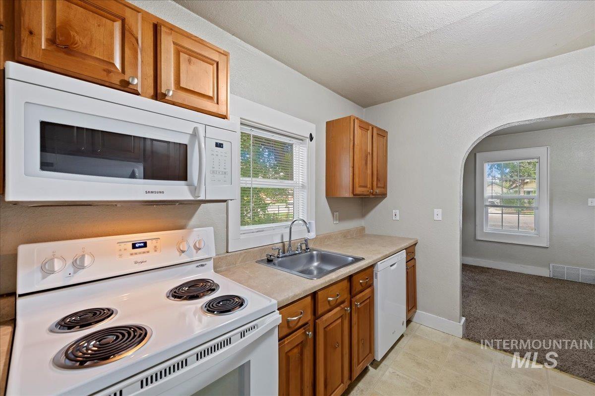 Kitchen with white appliances, arched walkways, brown cabinetry, light countertops, and a textured ceiling