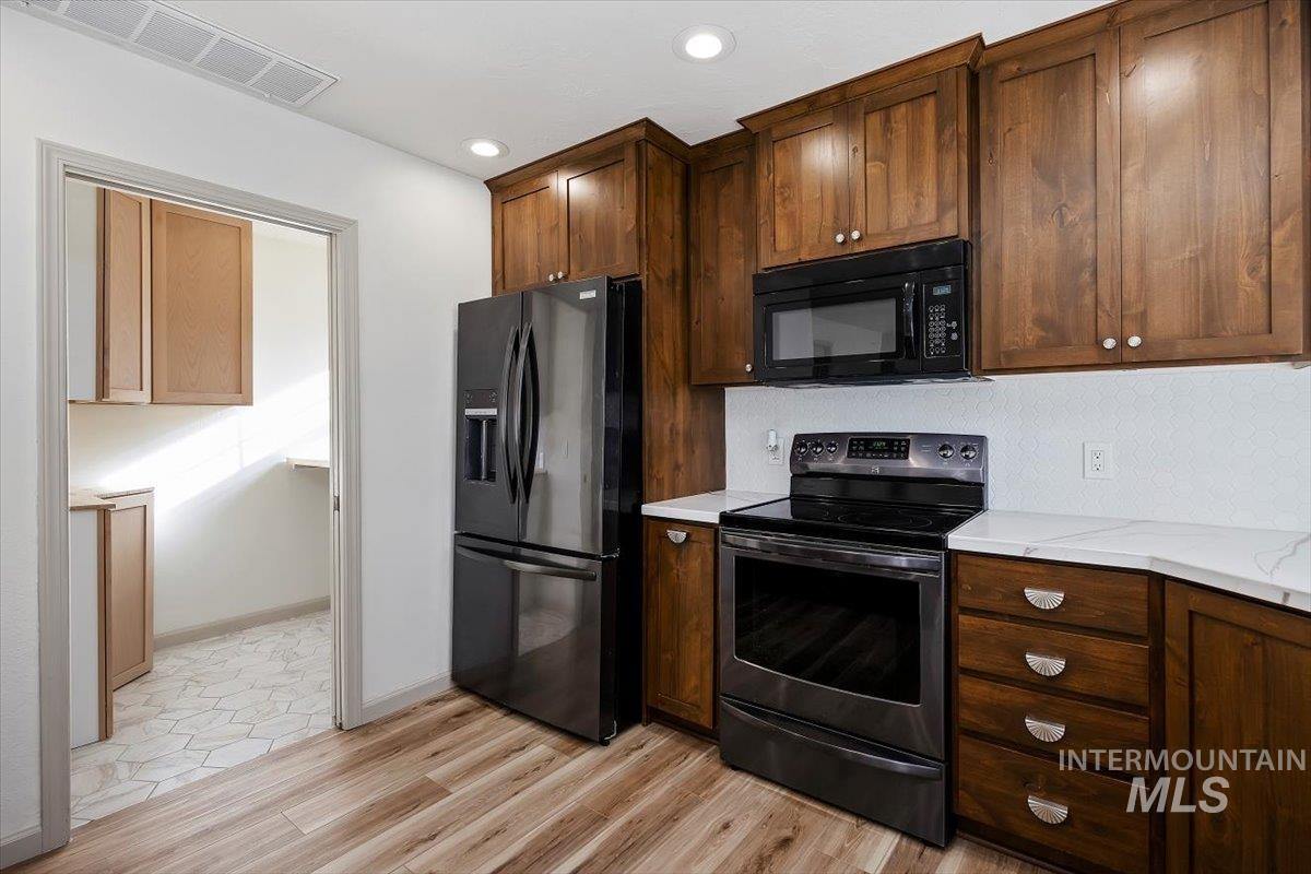 Kitchen with black appliances, dark brown cabinetry, light wood-type flooring, recessed lighting, and light stone counters