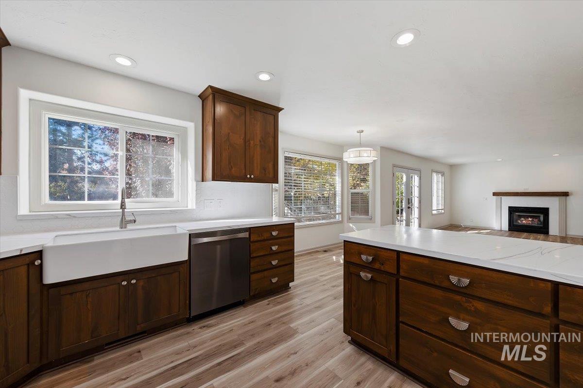 Kitchen with dark brown cabinets, a glass covered fireplace, light wood finished floors, stainless steel dishwasher, and hanging light fixtures