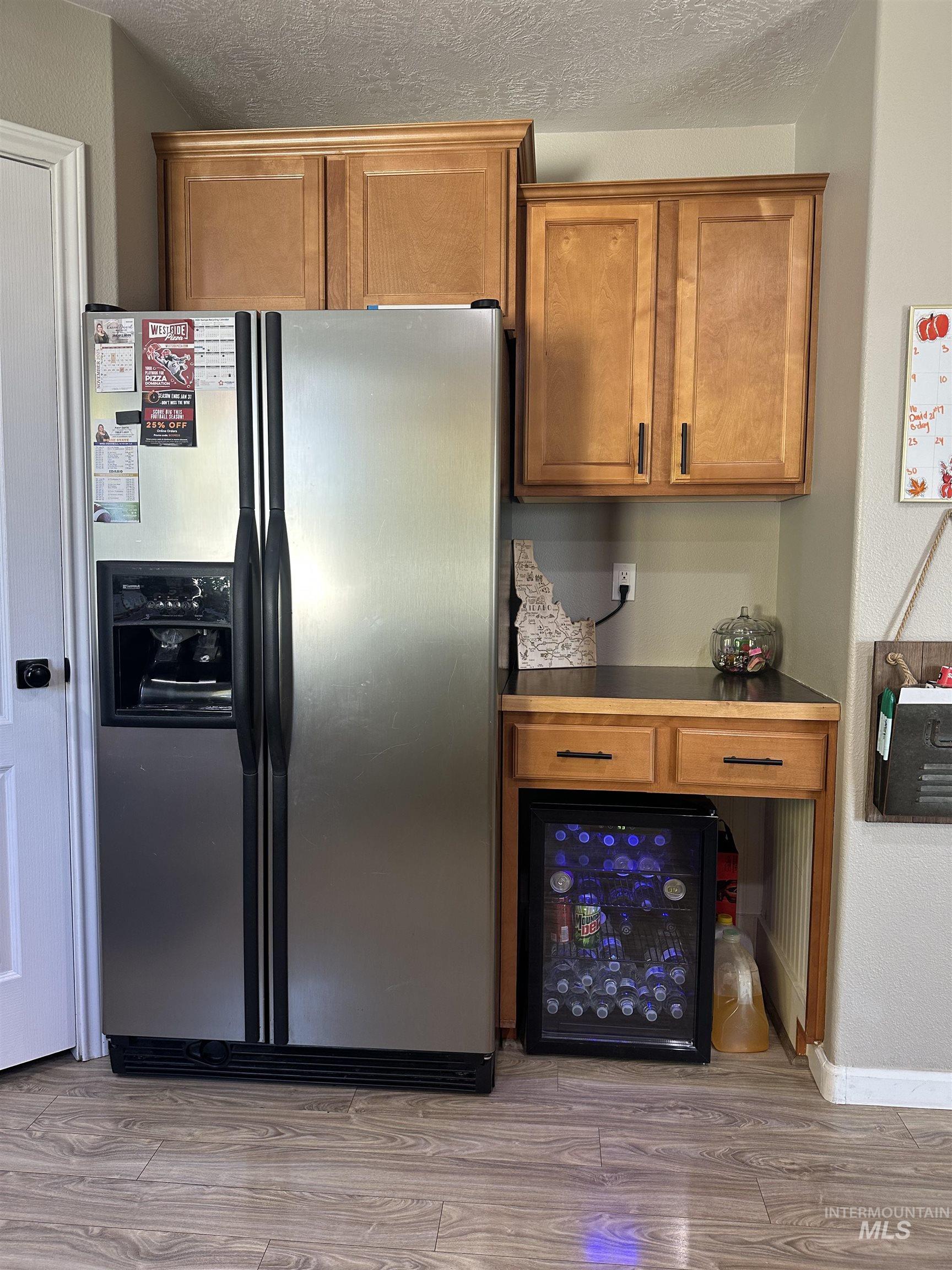Kitchen with stainless steel fridge with ice dispenser, brown cabinetry, light wood-type flooring, wine cooler, and a textured ceiling