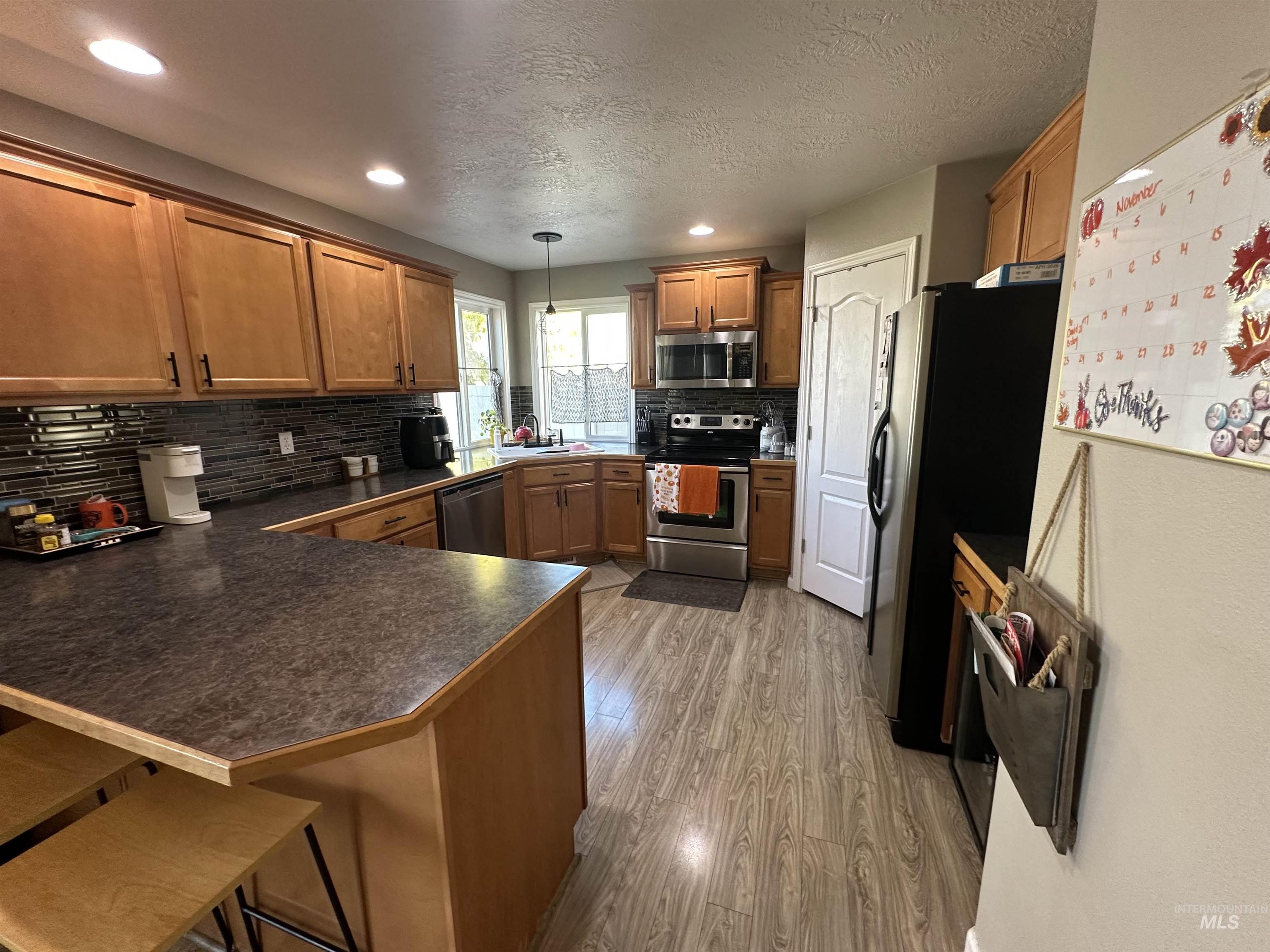 Kitchen with brown cabinets, dark countertops, stainless steel appliances, light wood-style floors, and decorative light fixtures
