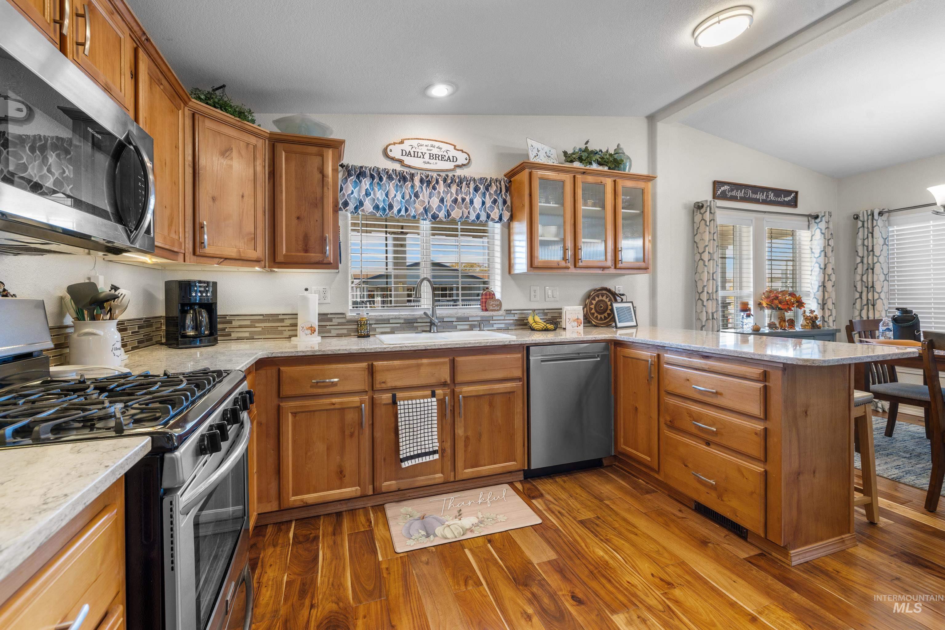 Kitchen with appliances with stainless steel finishes, lofted ceiling, a peninsula, brown cabinetry, and light stone countertops