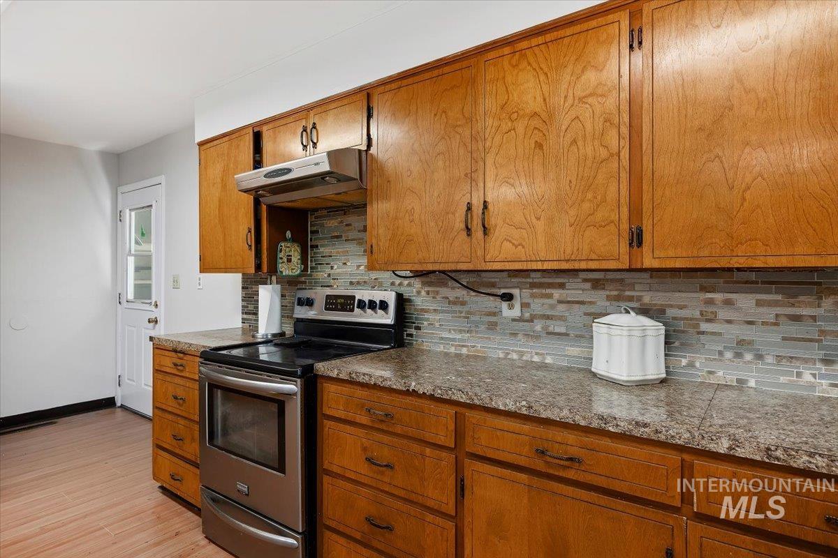 Kitchen featuring stainless steel electric range oven, decorative backsplash, brown cabinets, under cabinet range hood, and light wood-style floors
