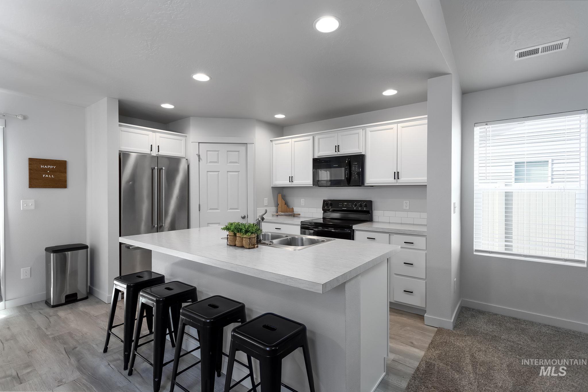 Kitchen featuring light countertops, white cabinets, a center island with sink, black appliances, and a kitchen bar