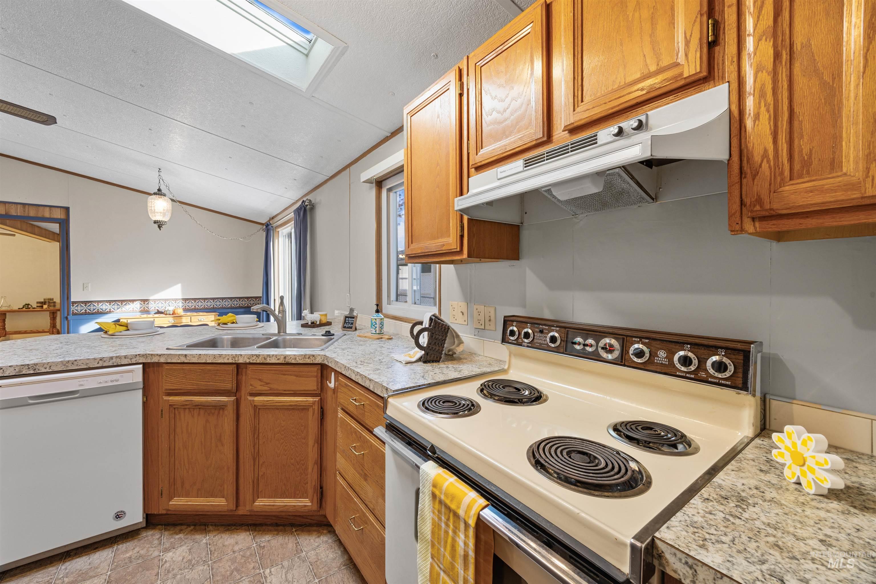 Kitchen featuring white appliances, brown cabinets, under cabinet range hood, light countertops, and vaulted ceiling