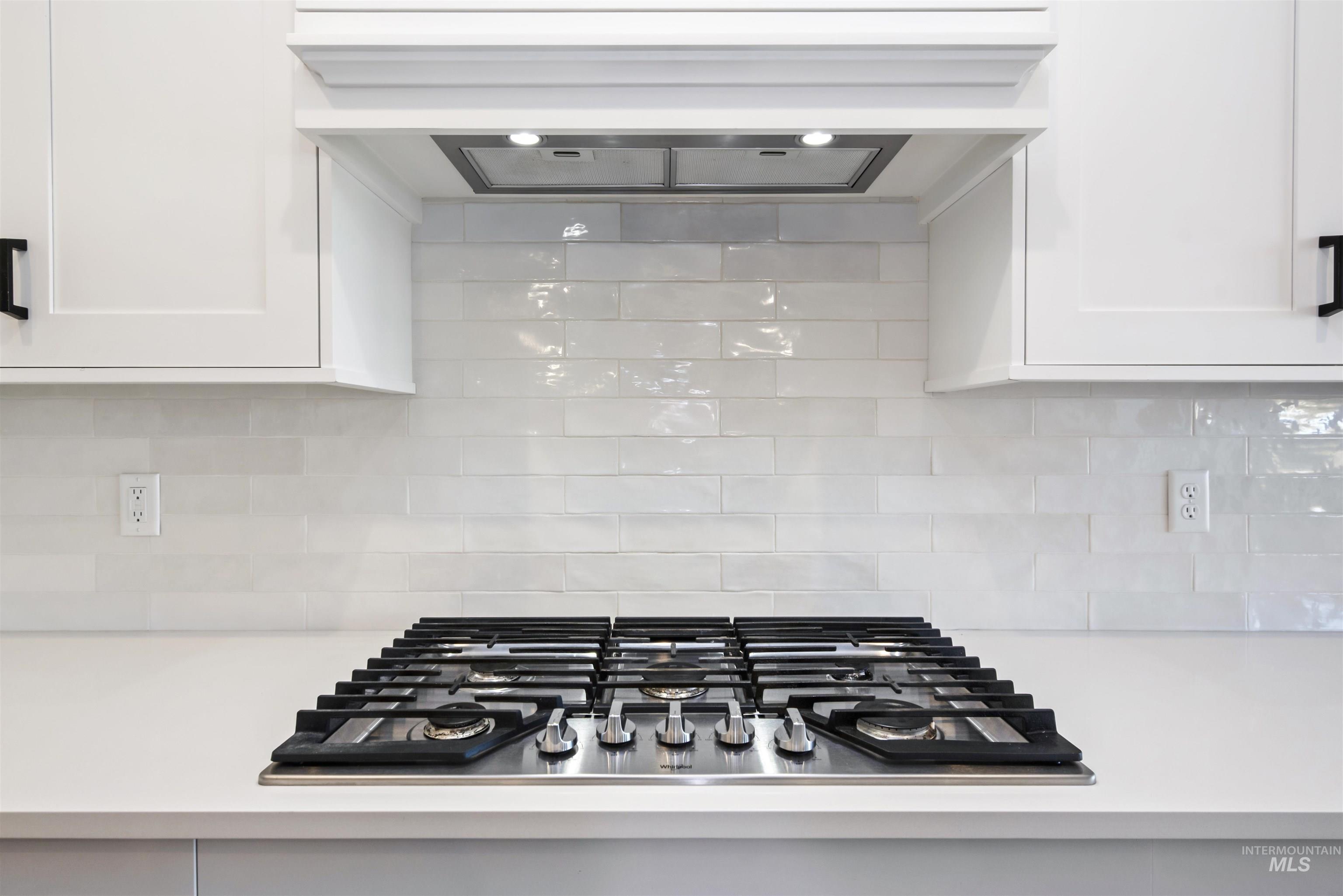 Kitchen view of exhaust hood, tasteful backsplash, stainless steel gas stovetop, white cabinetry, and light stone countertops