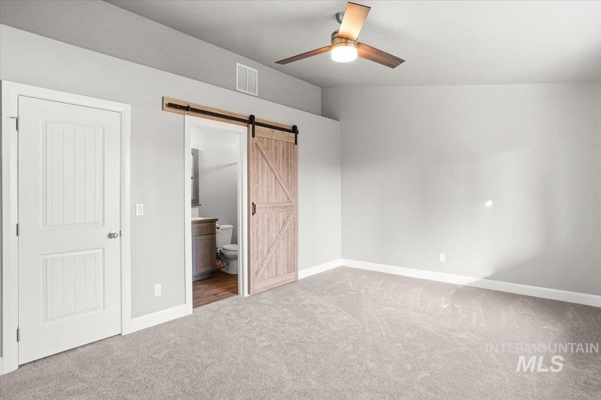 Unfurnished bedroom featuring ensuite bath, carpet flooring, a barn door, and ceiling fan