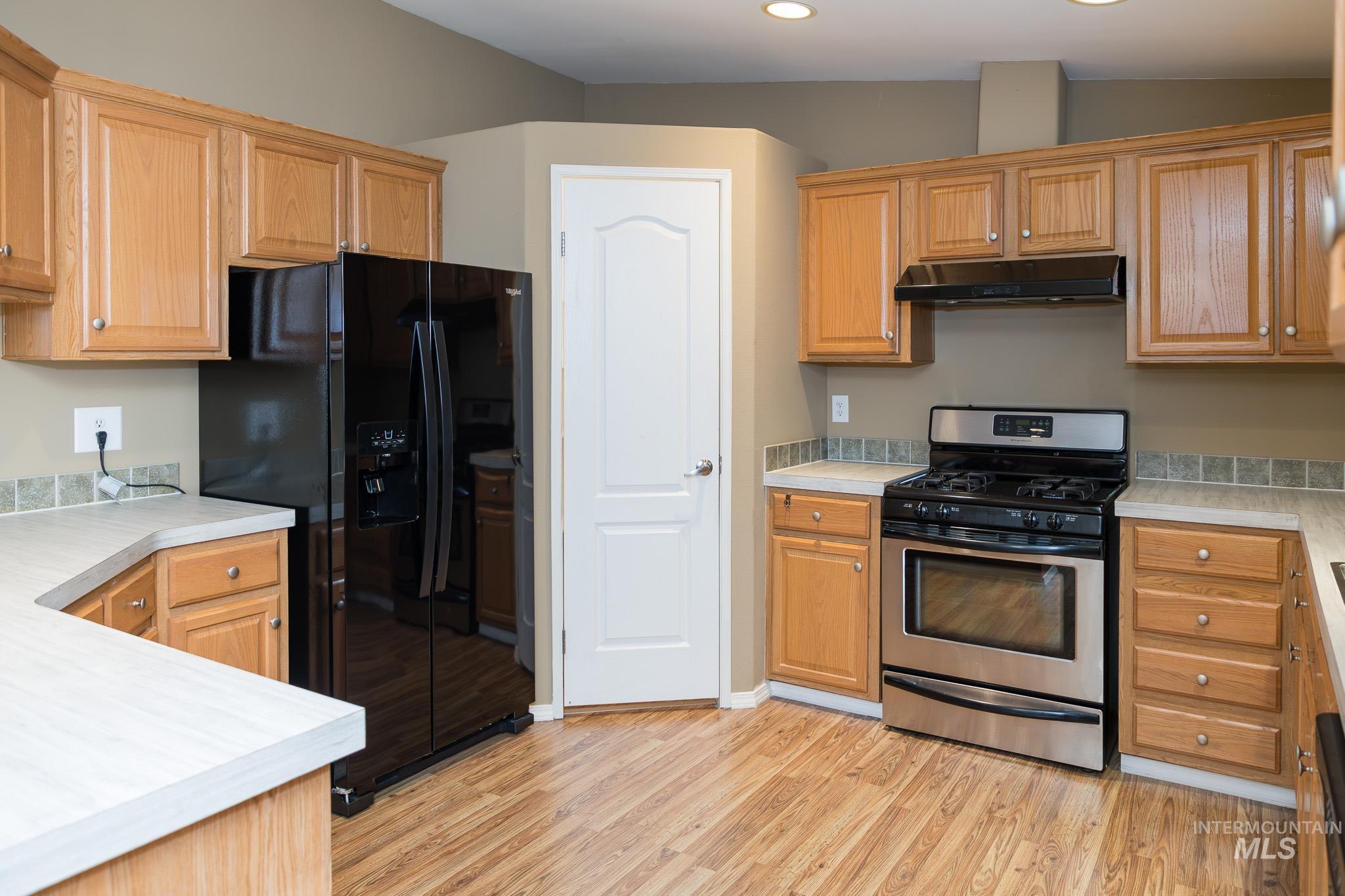 Kitchen with stainless steel gas stove, black refrigerator with ice dispenser, light countertops, light wood-type flooring, and under cabinet range hood