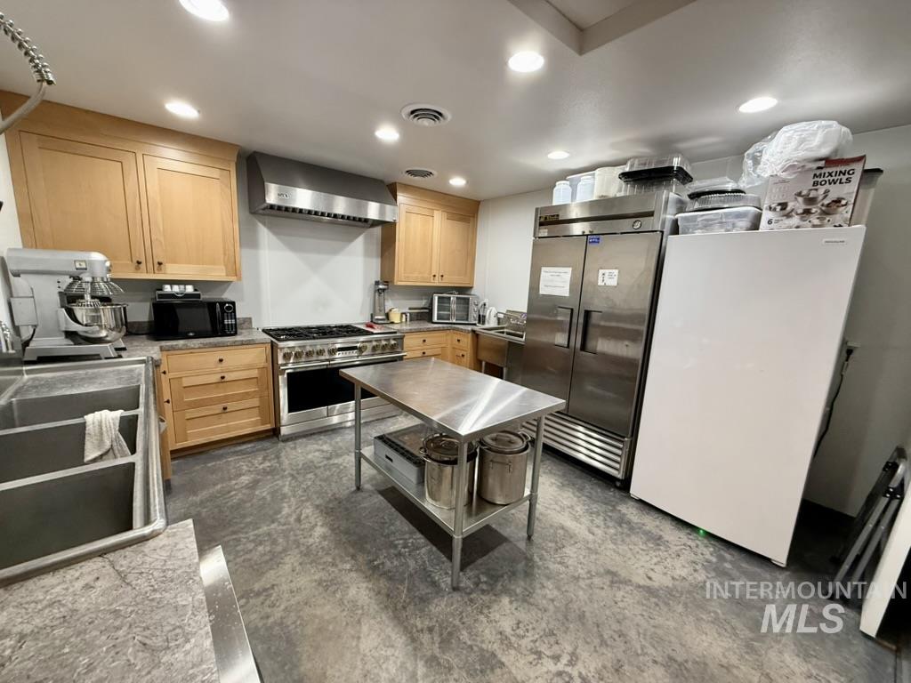 Kitchen featuring light brown cabinets, high end appliances, recessed lighting, wall chimney exhaust hood, and concrete flooring