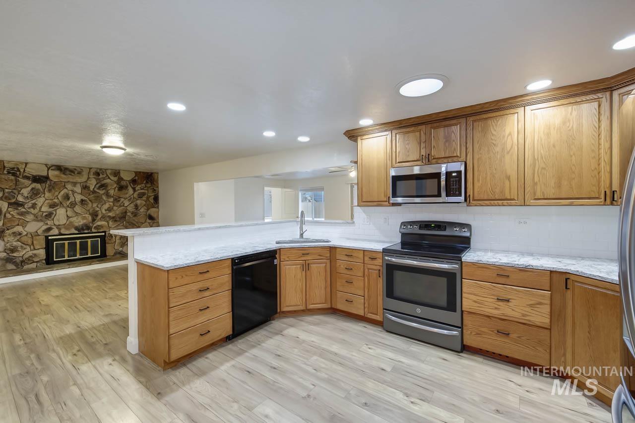 Kitchen featuring appliances with stainless steel finishes, a peninsula, brown cabinetry, a stone fireplace, and light wood-type flooring