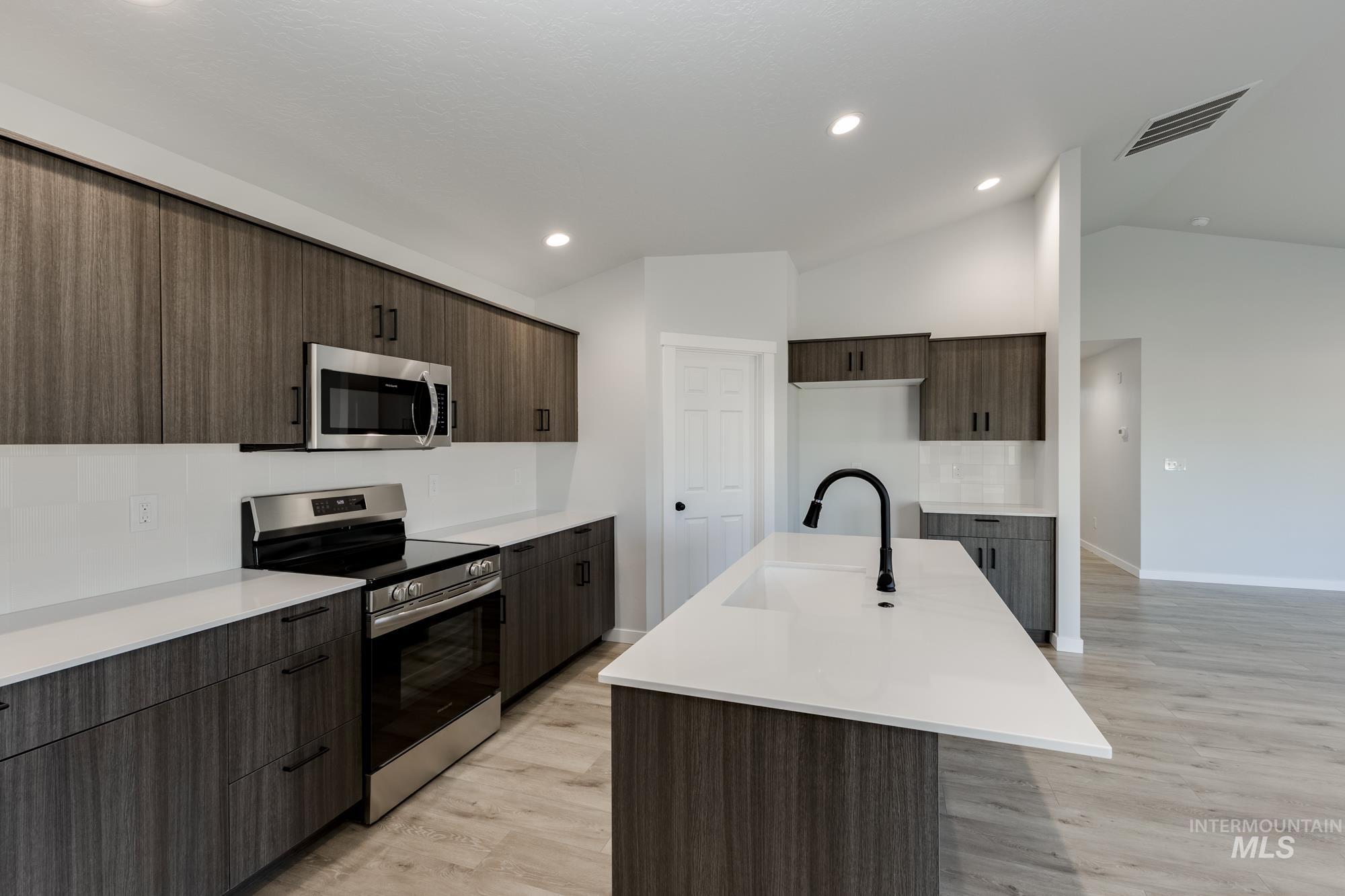 Kitchen with stainless steel appliances, light wood-style flooring, a kitchen island with sink, vaulted ceiling, and dark brown cabinetry