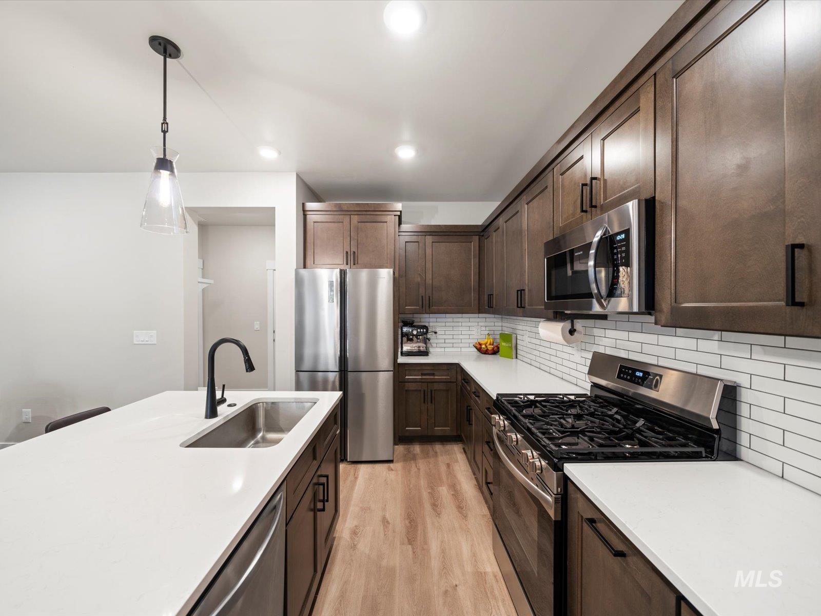 Kitchen with stainless steel appliances, dark brown cabinets, hanging light fixtures, backsplash, and light wood-style flooring