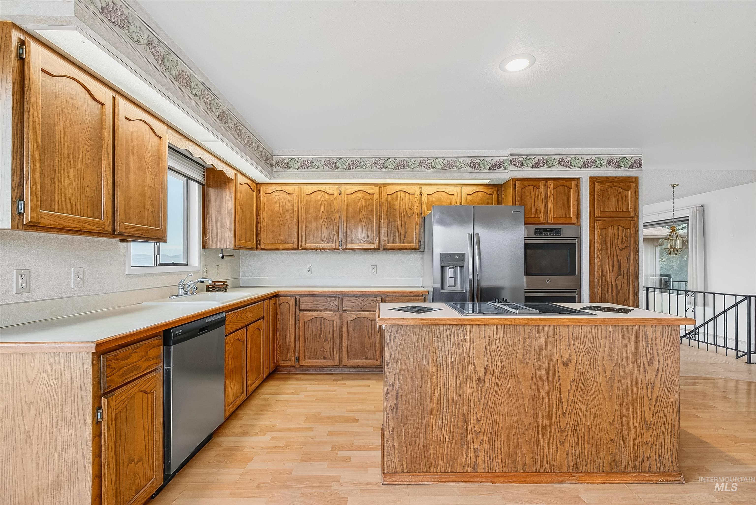 Kitchen with brown cabinetry, stainless steel appliances, light wood-style flooring, a center island, and light countertops