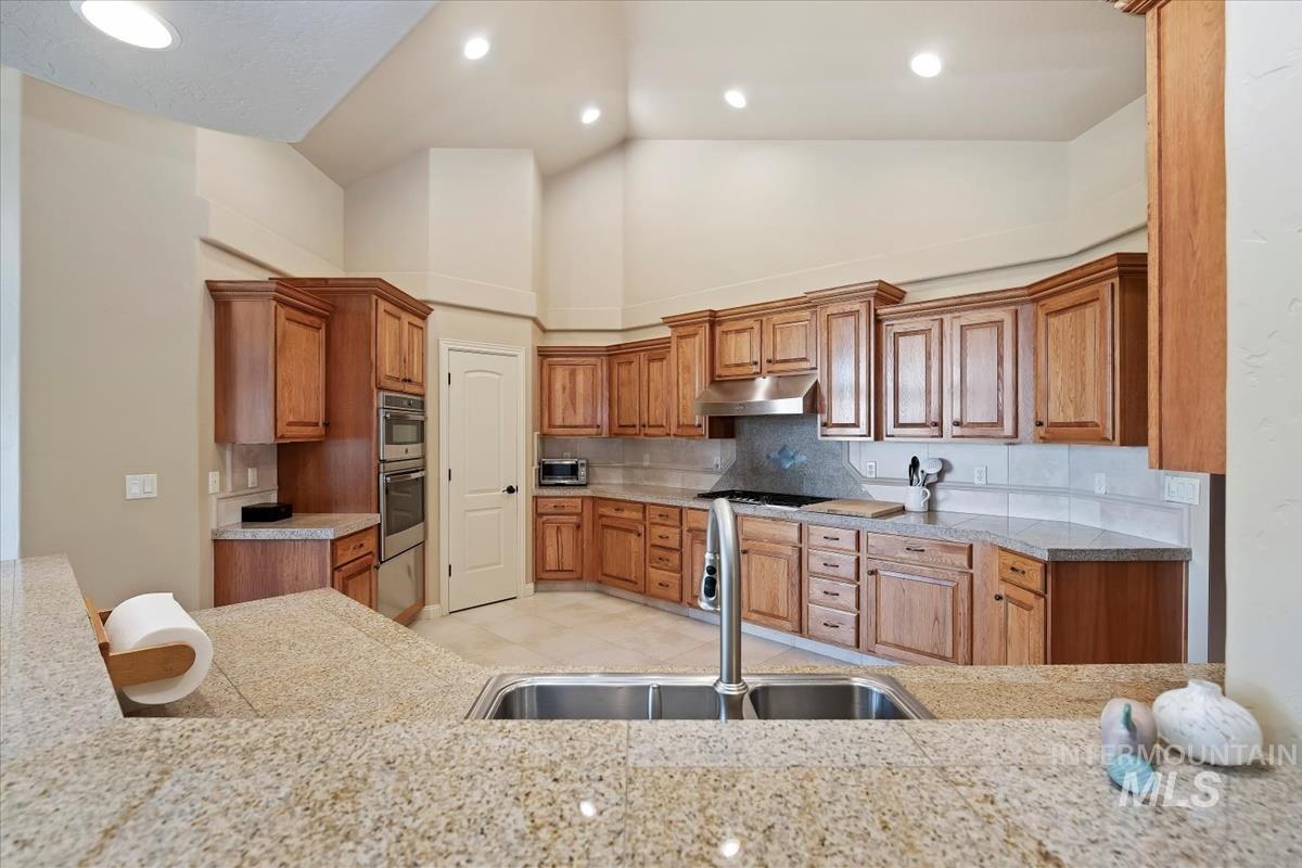 Kitchen featuring brown cabinets, backsplash, under cabinet range hood, high vaulted ceiling, and recessed lighting