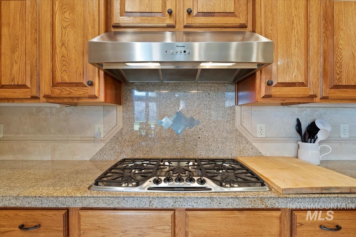 Kitchen with under cabinet range hood, backsplash, stainless steel gas cooktop, and brown cabinetry