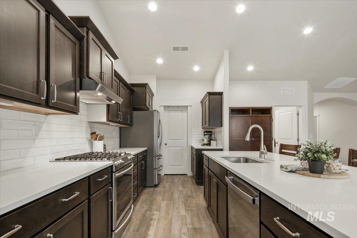 Kitchen with dark brown cabinets, stainless steel appliances, light wood-style flooring, tasteful backsplash, and under cabinet range hood