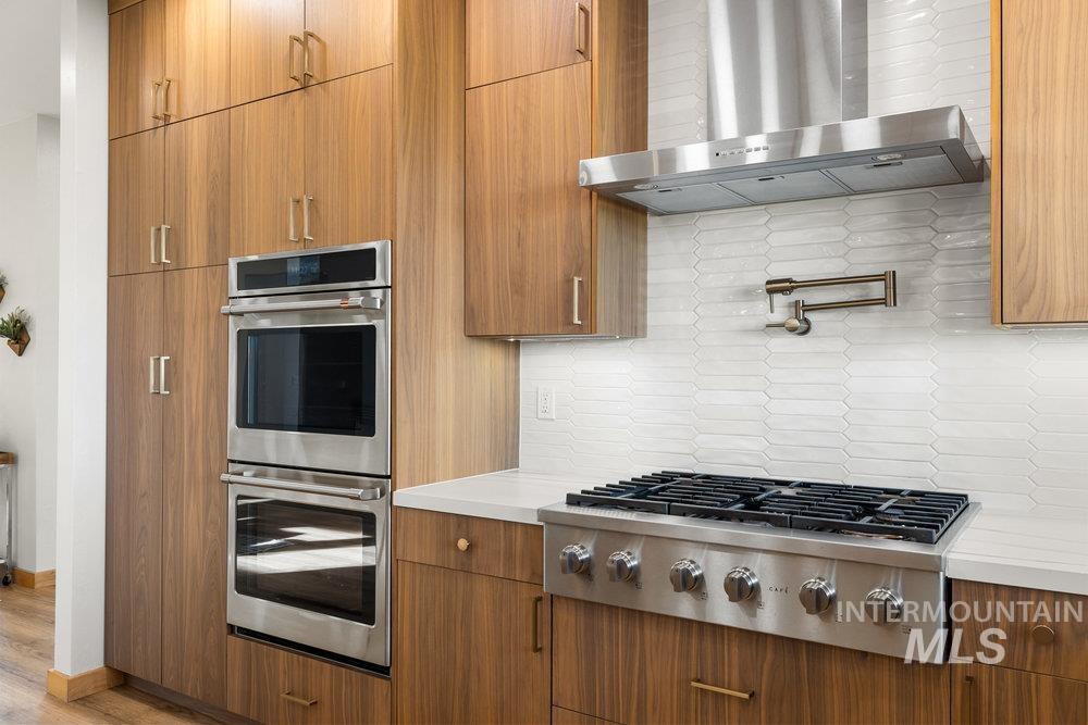 Kitchen with appliances with stainless steel finishes, wall chimney exhaust hood, brown cabinetry, and tasteful backsplash
