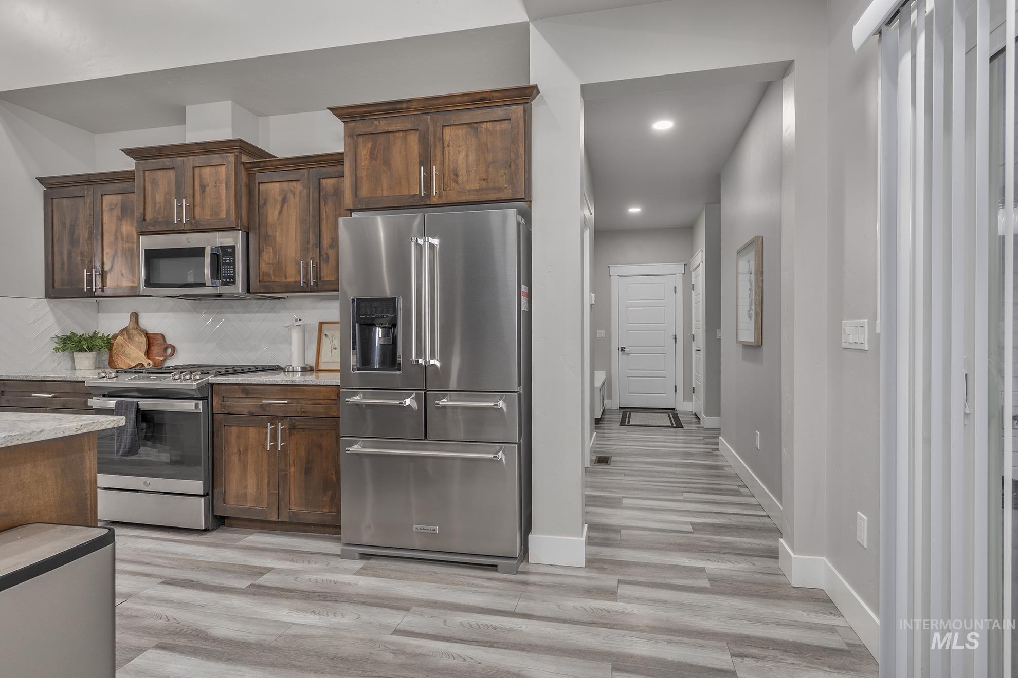 Kitchen featuring appliances with stainless steel finishes, dark brown cabinetry, decorative backsplash, light stone countertops, and light wood-style flooring