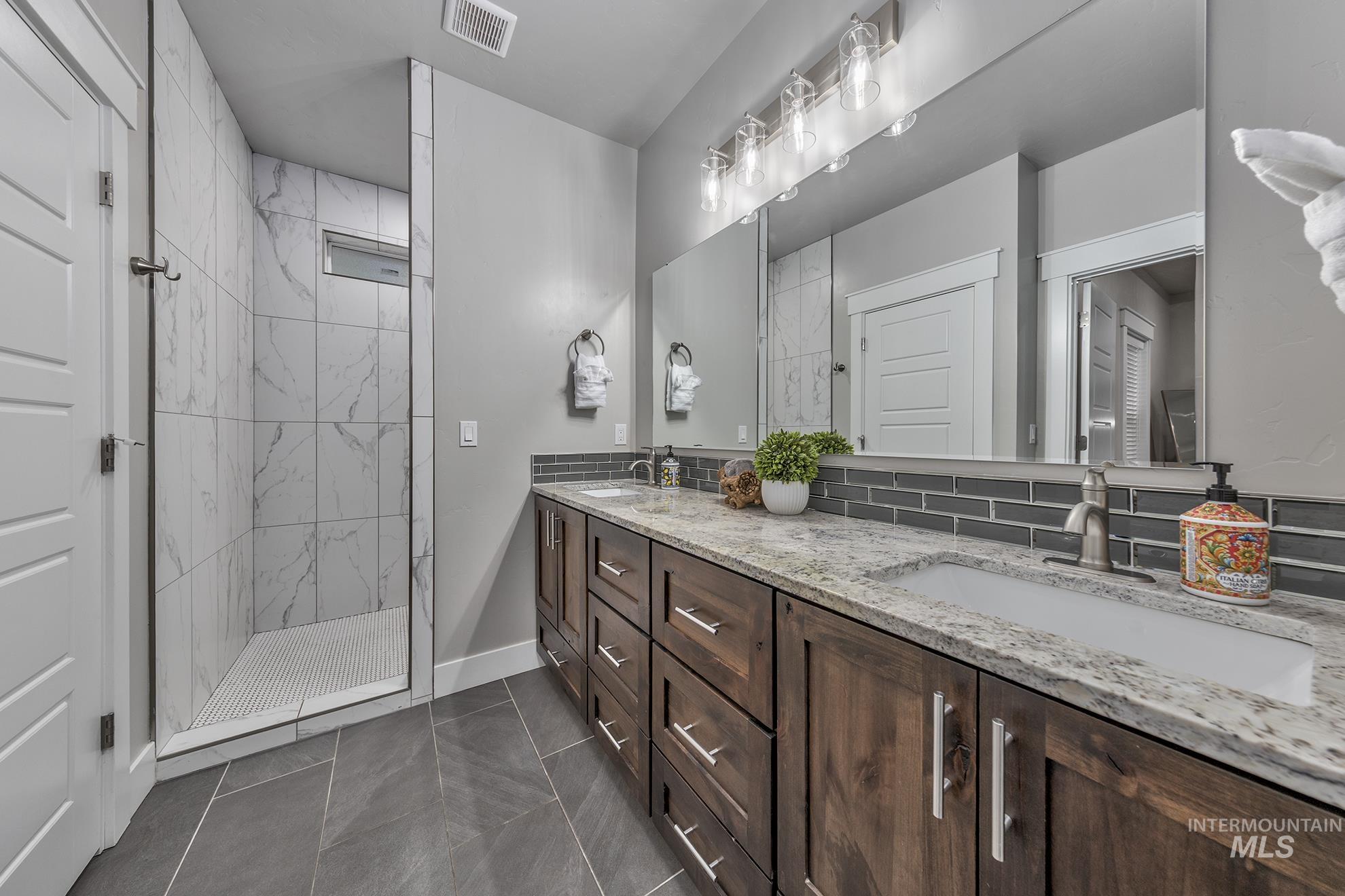 Bathroom with double vanity, a tile shower, decorative backsplash, and dark tile patterned flooring