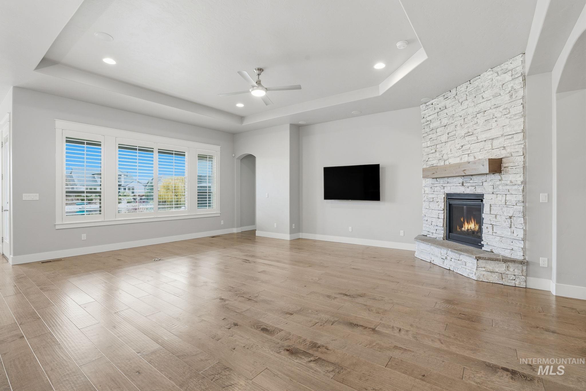Unfurnished living room with arched walkways, light wood finished floors, a stone fireplace, a raised ceiling, and recessed lighting